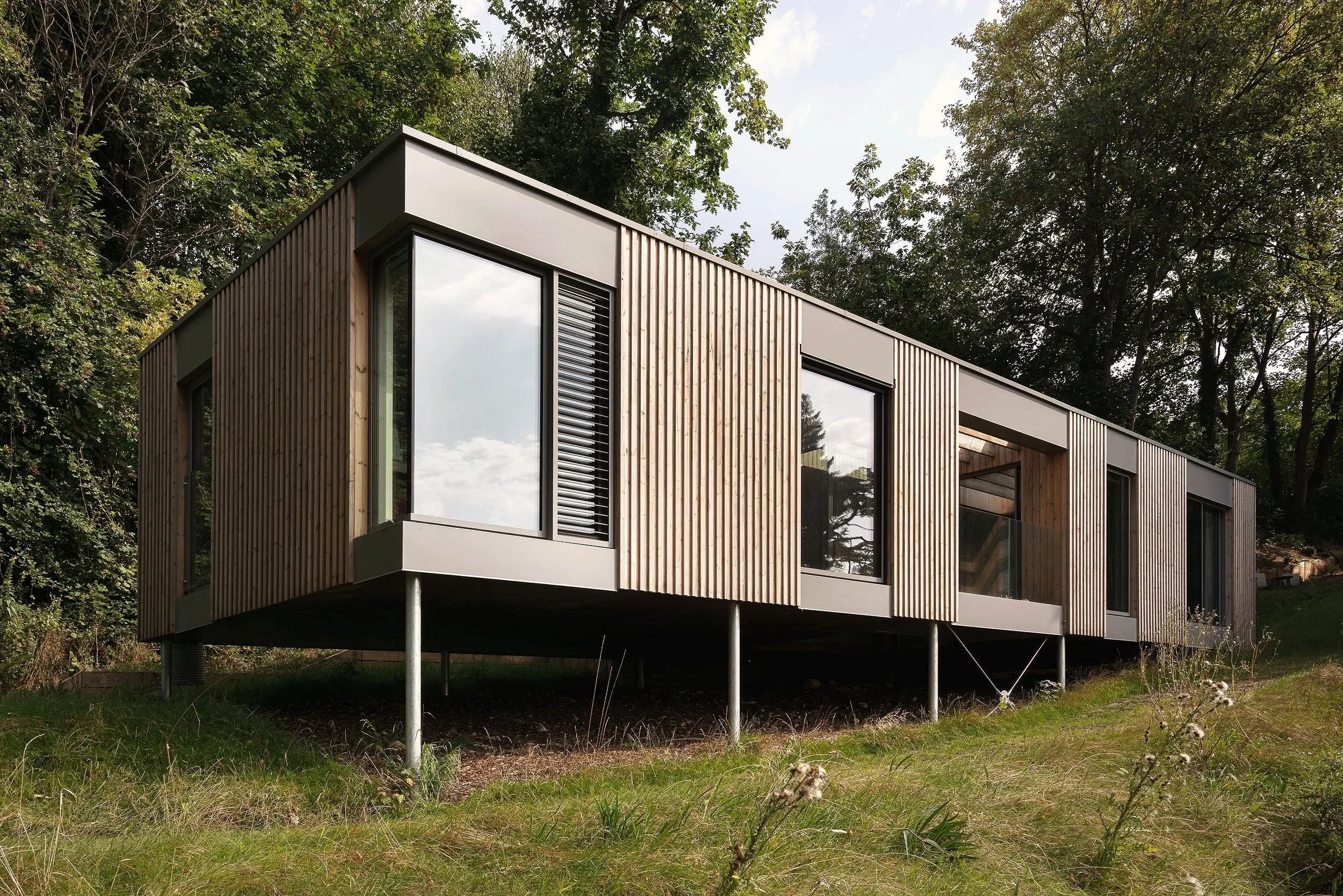 Modern house with wooden panel exterior, elevated on metal stilts, surrounded by trees and grass.