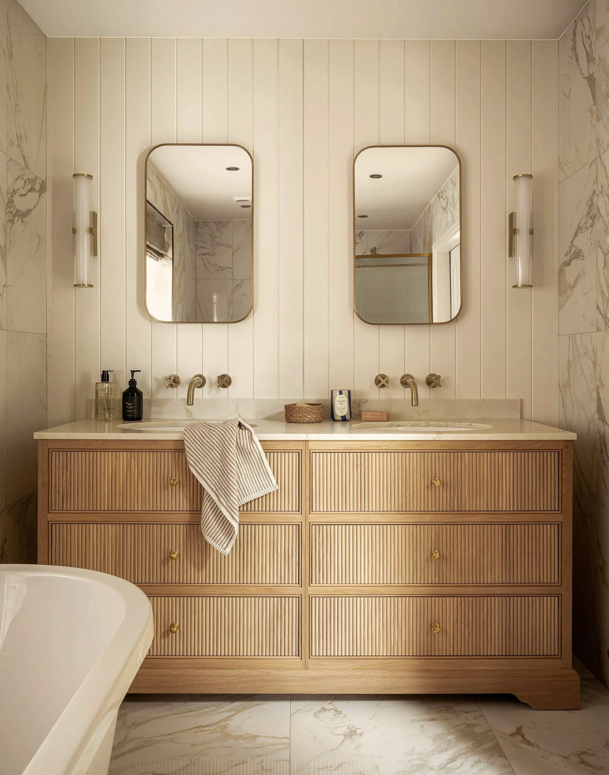 A dual vanity bathroom with two oval mirrors above a light wood cabinet with six drawers, a marble countertop, two wall-mounted sinks, and wall-mounted light fixtures.