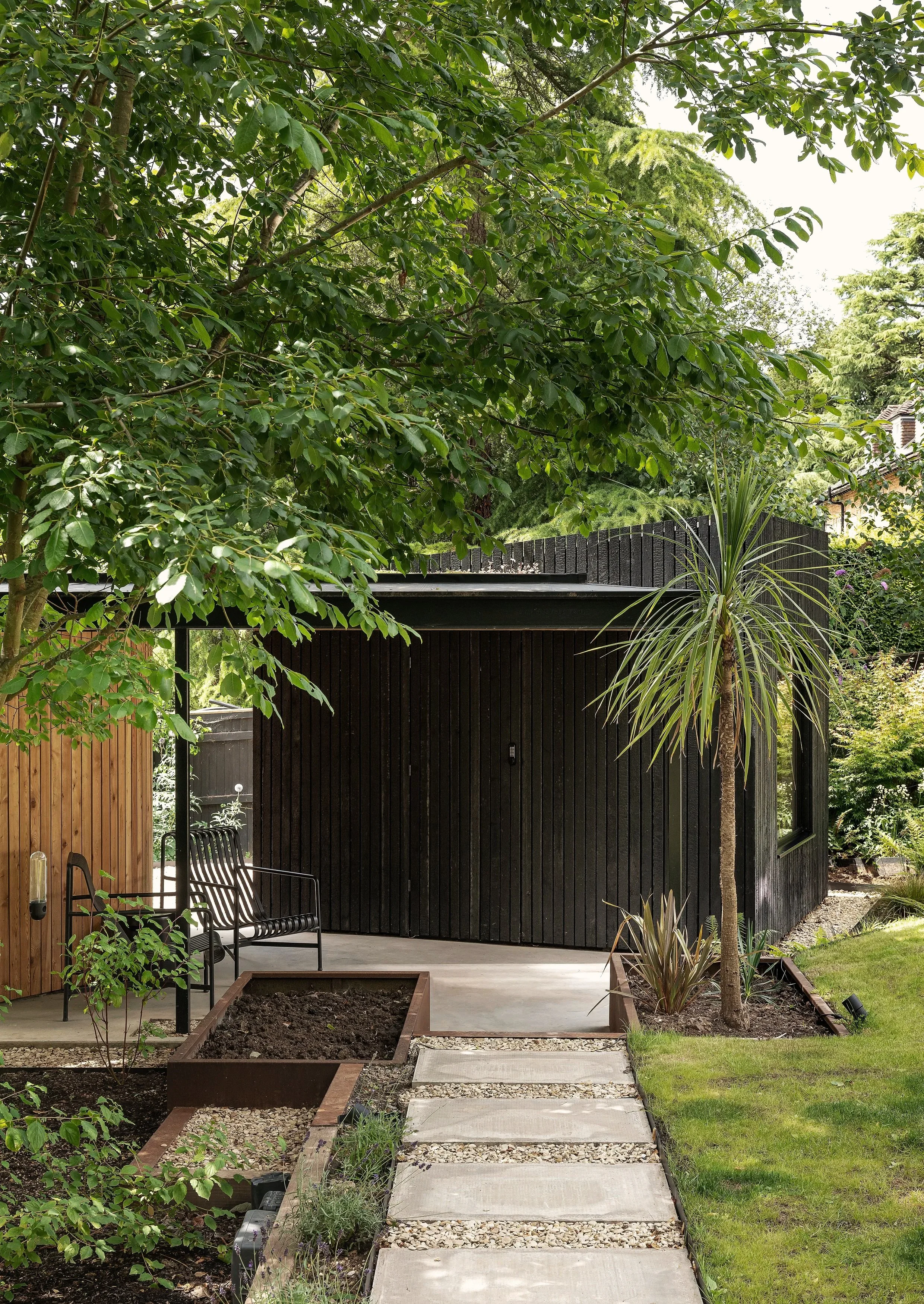 Modern black and wood residential building with a small patio, surrounded by green trees and plants, including a tall palm-like tree, with a concrete pathway leading to the entrance.