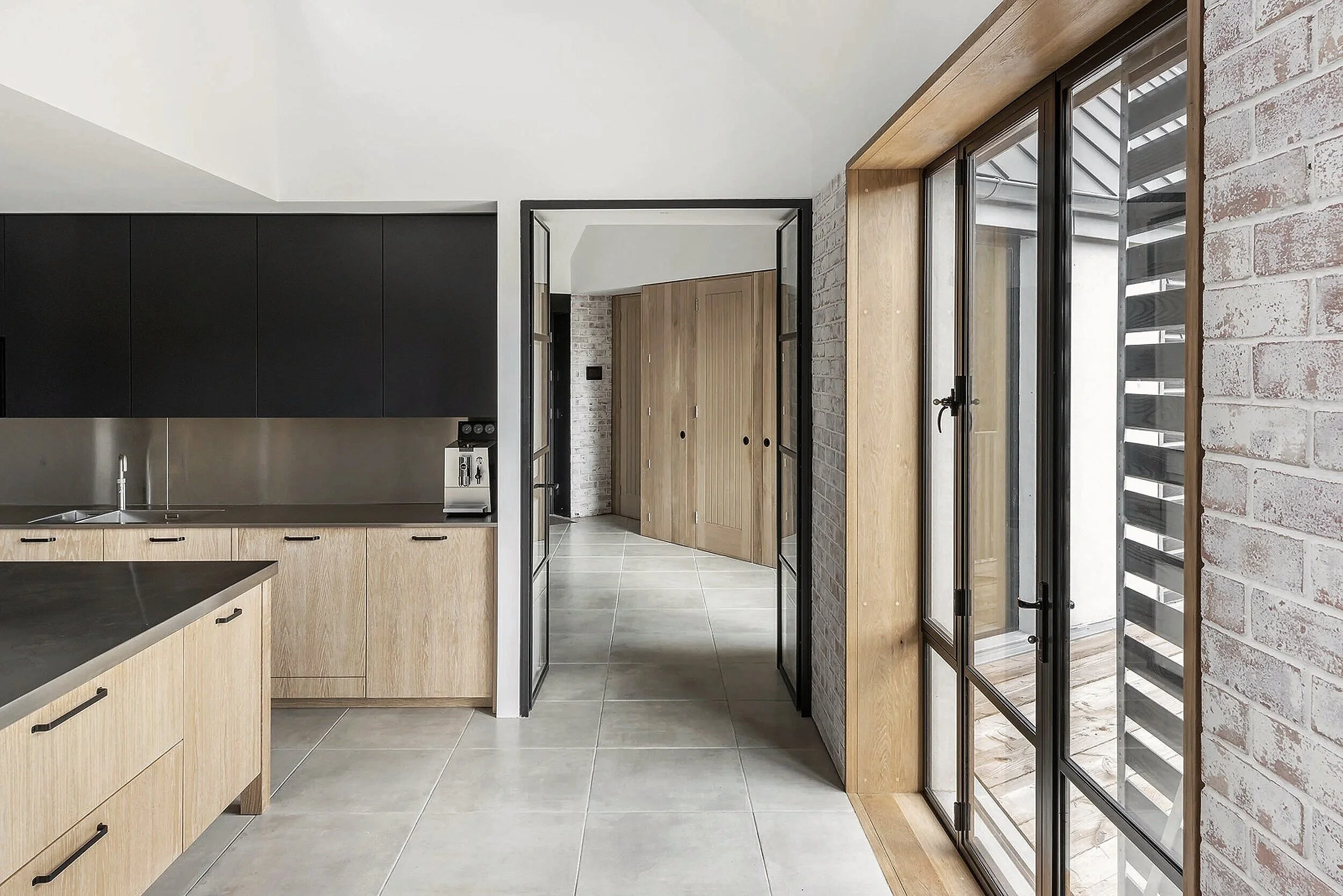 Modern kitchen with black cabinets, light wood lower cabinets, a stainless steel countertop, and large glass doors with black metal frames leading to an outdoor space, exposed white brick wall on the right.