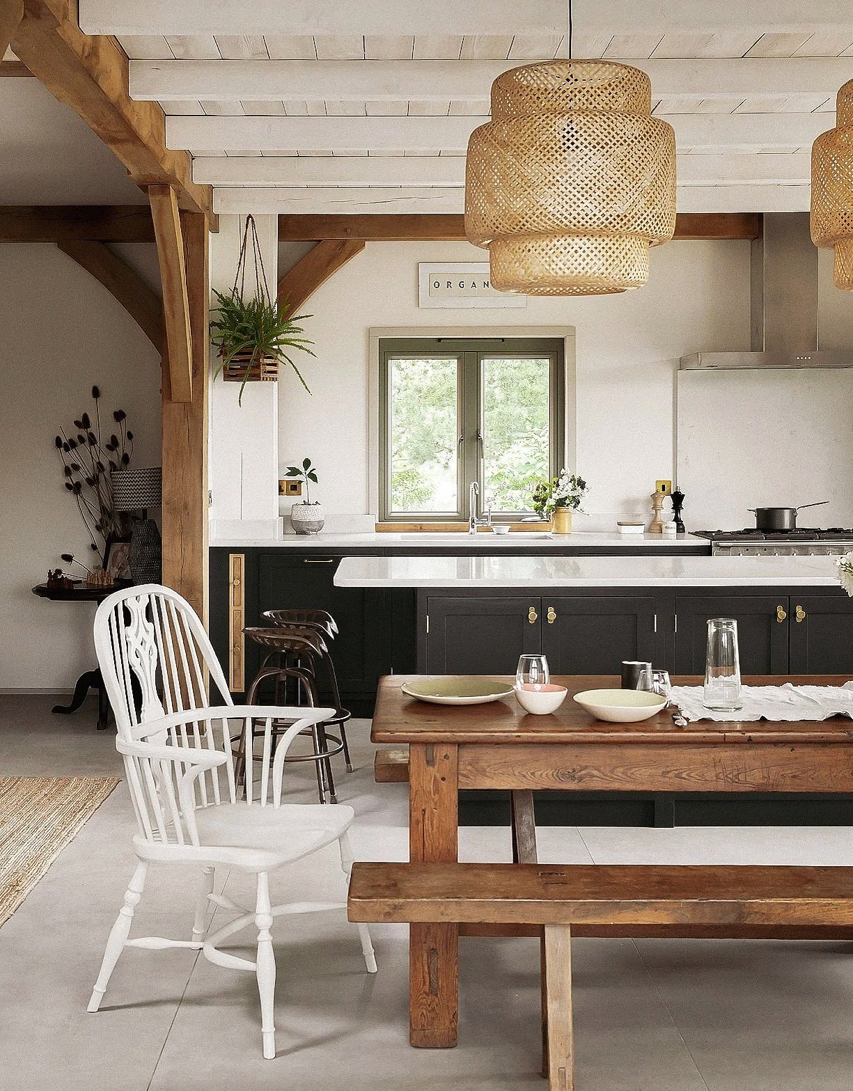 A cozy, modern kitchen with a white ceiling, black cabinets, and a large window above the sink. There are woven rattan pendant lights hanging from the ceiling. A wooden dining table has bowls and a glass pitcher, accompanied by a white wooden chair a