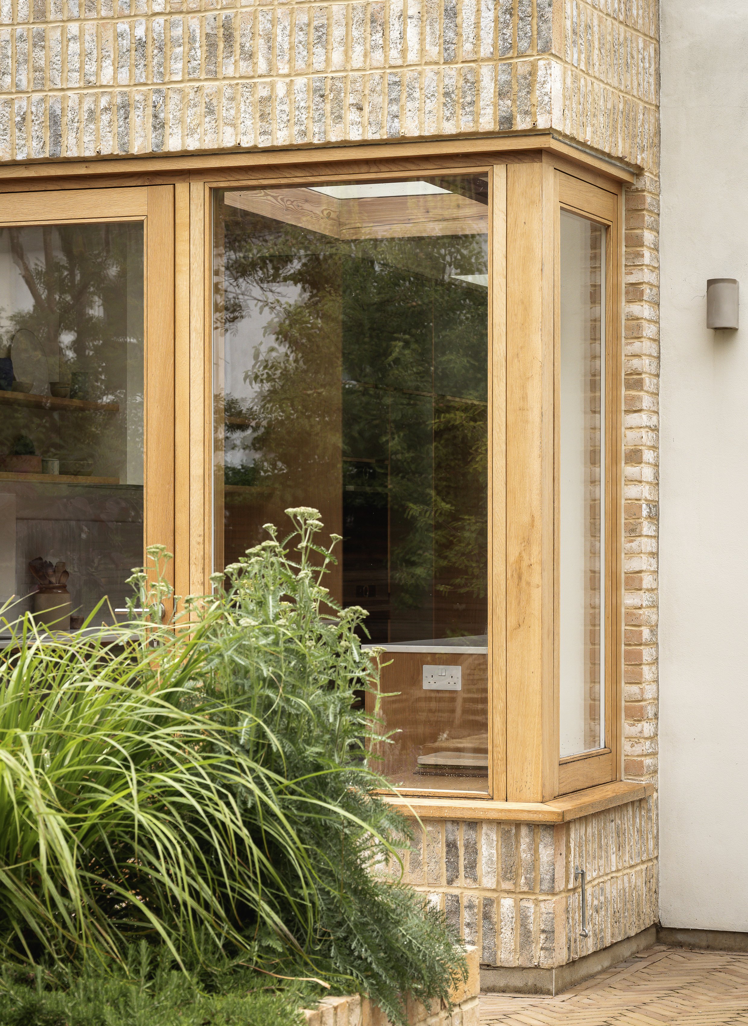 Photo of a house exterior with a large bay window framed in natural wood, surrounded by green plants and brickwork.