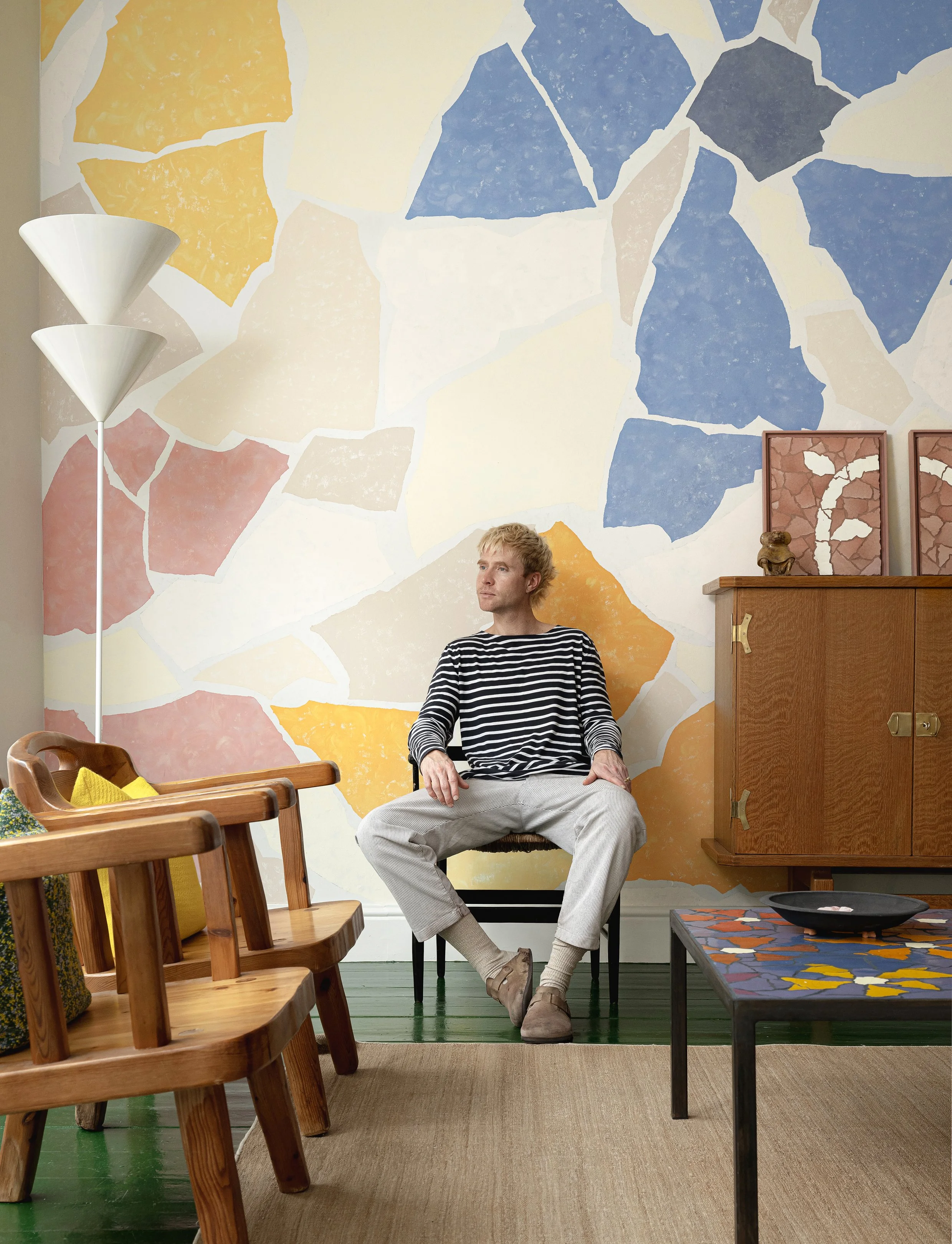 A man with blond hair wearing a striped shirt and light-colored pants sitting on a chair in a colorful room with a large abstract mural on the wall, wooden furniture, and a black coffee table.