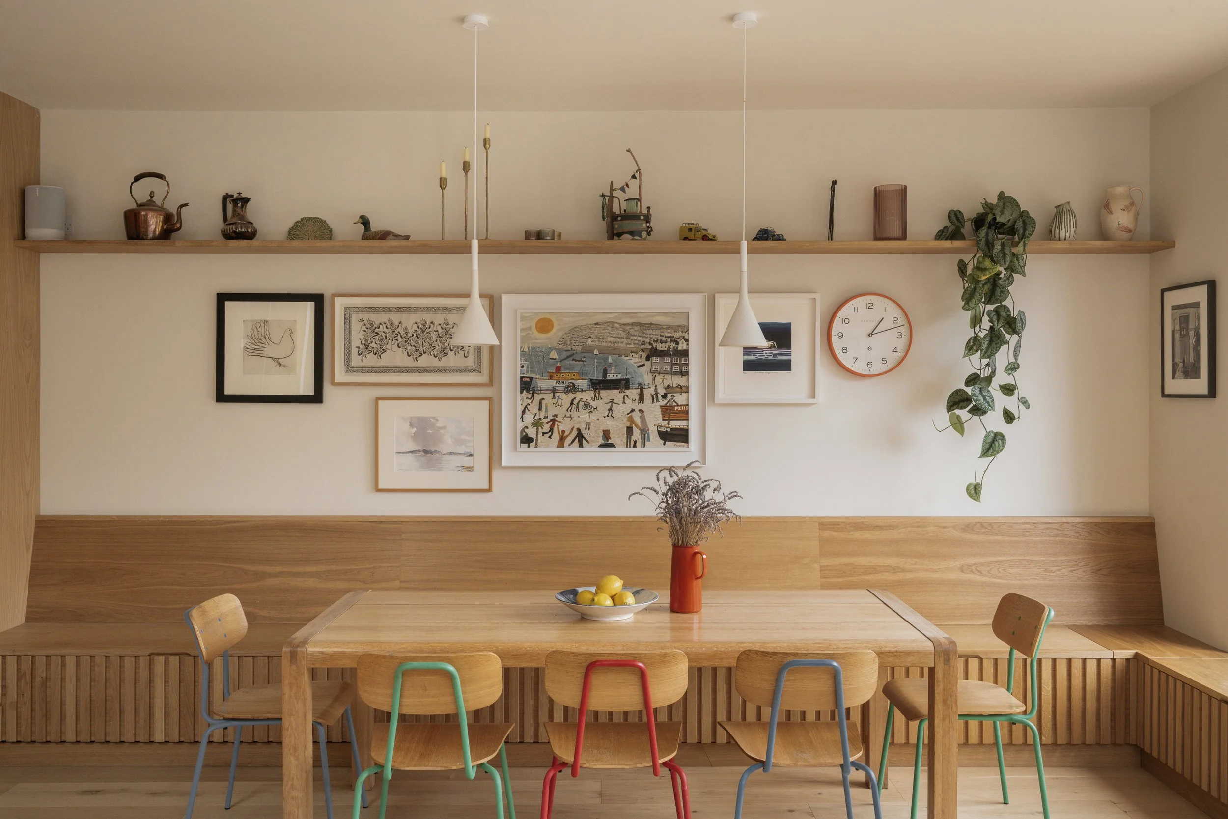 A dining area with a wooden table surrounded by five chairs with colorful legs. On the table, there is a gray bowl with lemons and a red vase with dried flowers. The wall behind has a gallery of framed art and a wooden shelf with decorative objects a