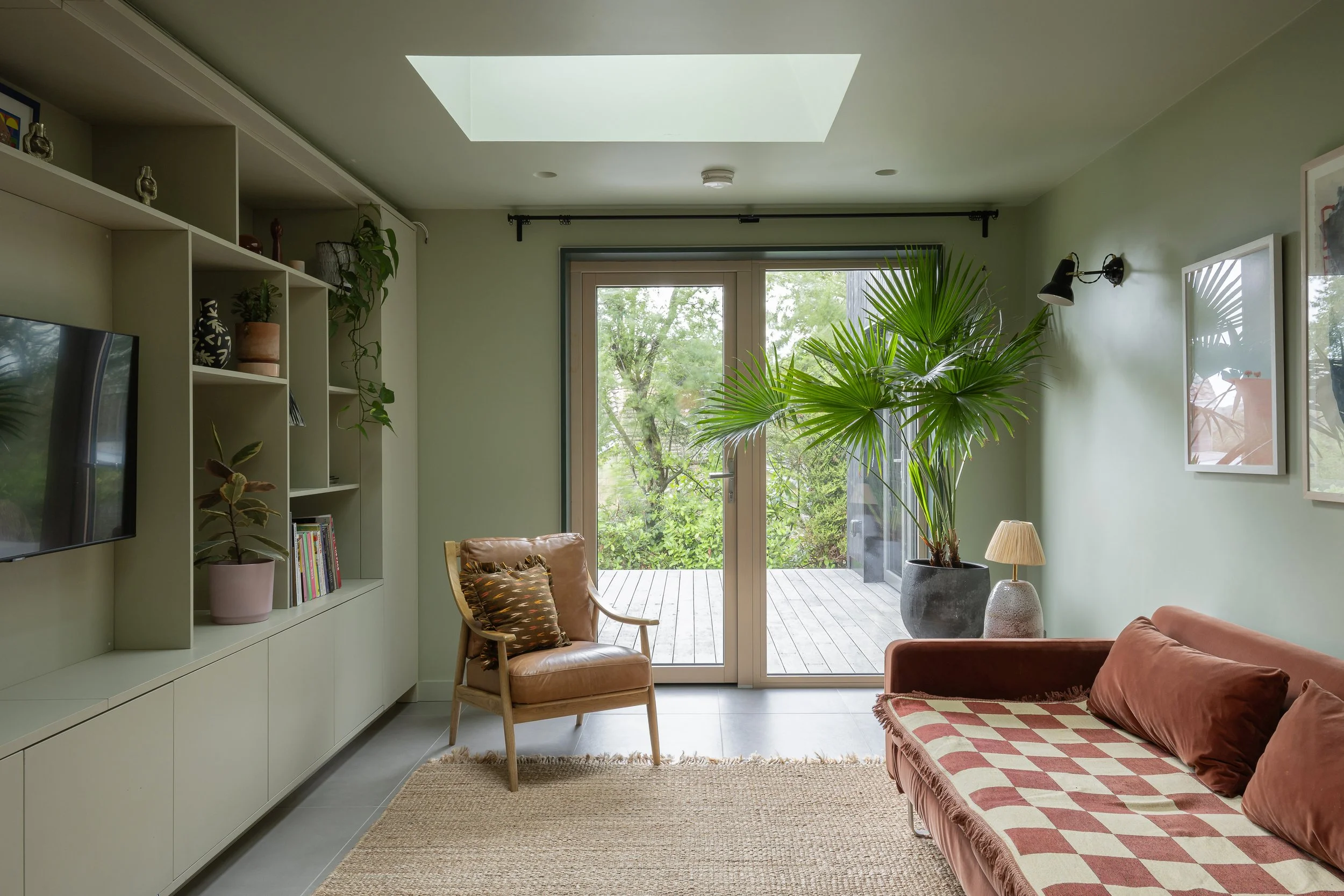 Living room with a large potted plant near glass sliding door, beige armchair with pillow, rust-colored sofa with pillows, green walls, built-in shelves with decor, skylight in ceiling, wall-mounted lamps, framed art, and outdoor deck visible through