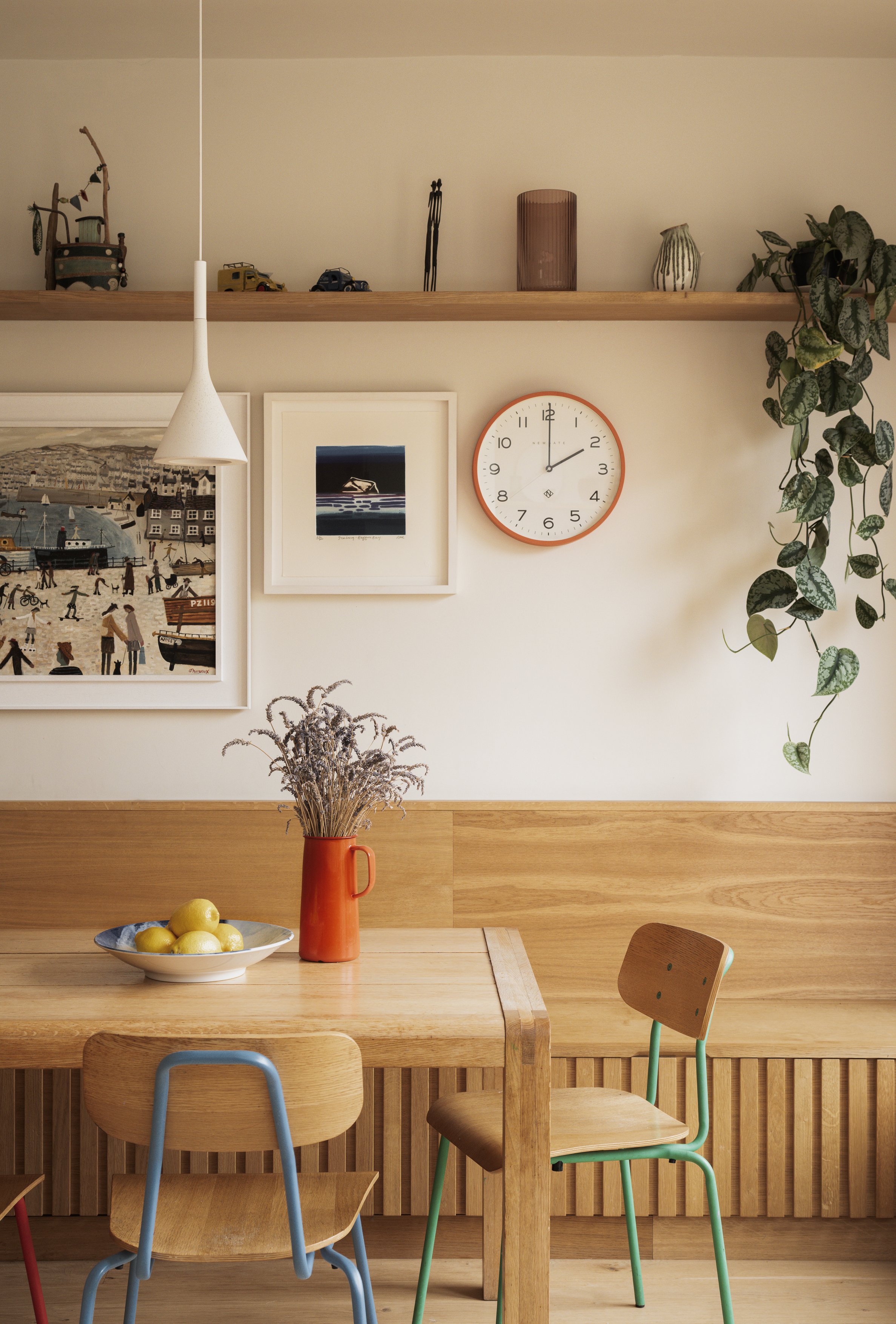 Interior of a wooden dining area with a wooden table, a bowl of lemons, and colorful chairs. The wall behind features framed artwork, a round wall clock, a hanging pendant light, and a shelf with decorative items and a plant.
