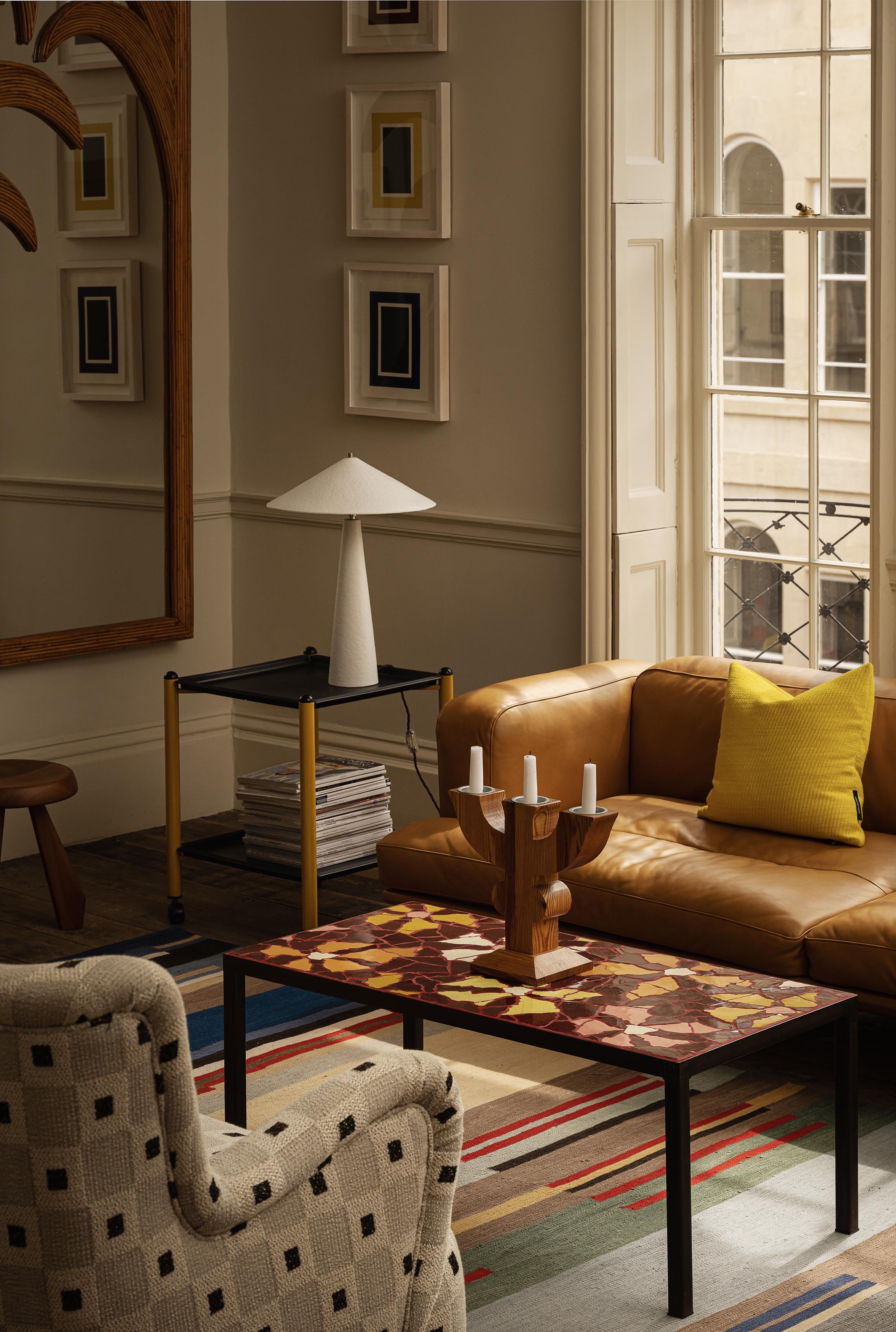 Living room with beige walls, a large window, a leather sofa with a yellow pillow, a patterned armchair, a colorful rug, a coffee table with a decorative wooden candle holder, a side table with a white lamp, and a wall with framed artwork.