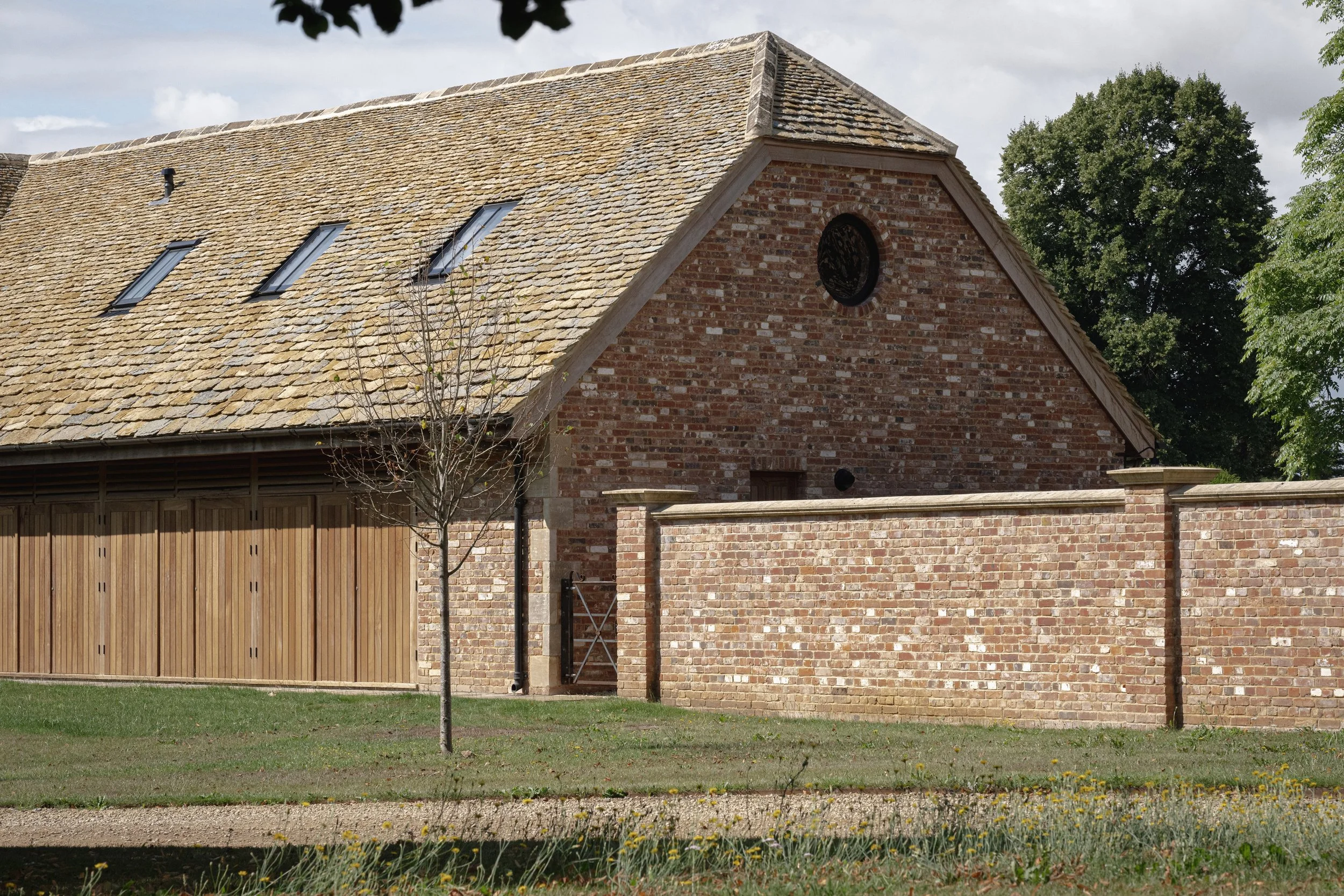 A brick building with a steep roof covered in tan shingles, a small leafless tree in front, and a brick wall with a gate. There are four skylights on the roof and a round window near the peak of the building.