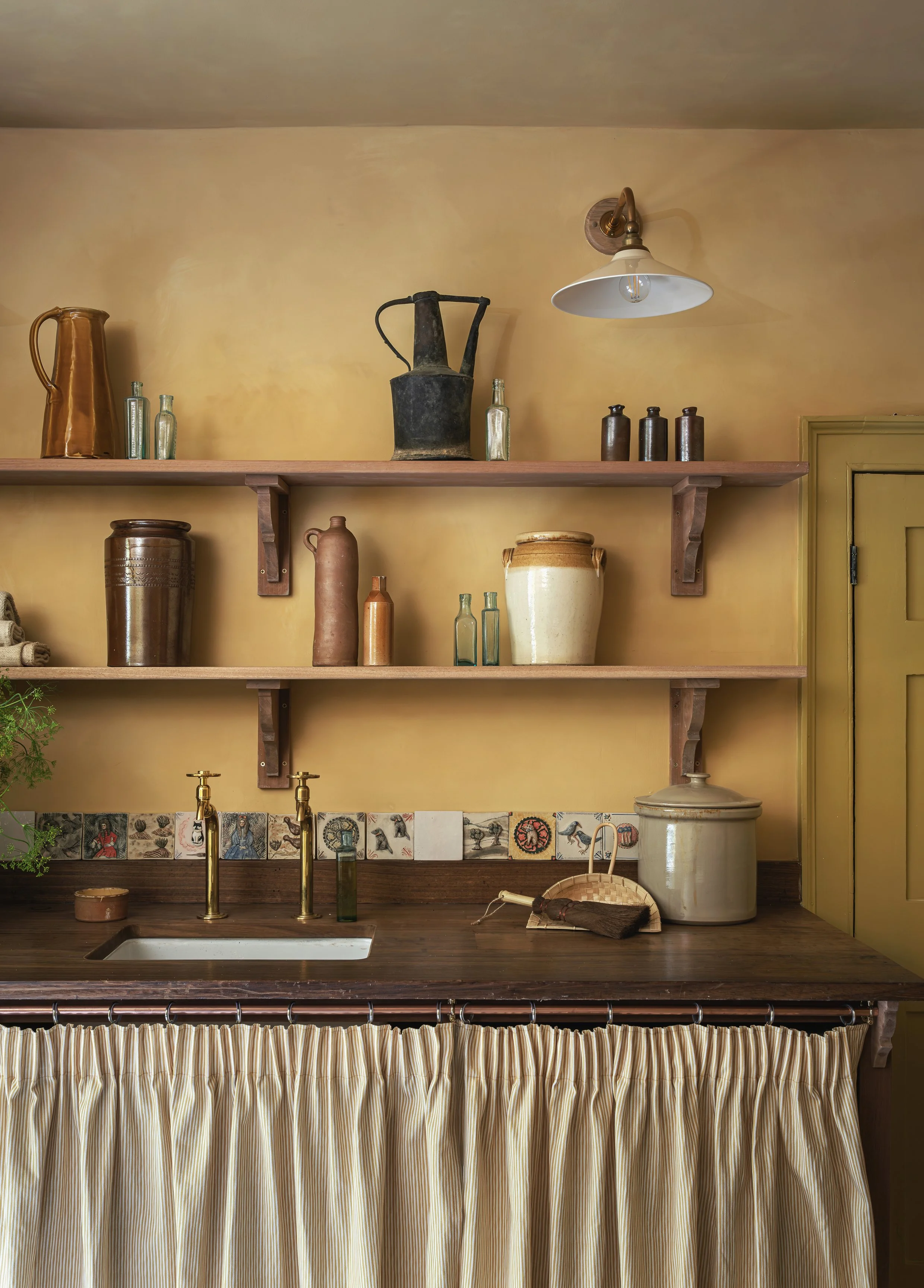 Interior of a rustic kitchen with open wooden shelves holding vintage containers and bottles above a wooden countertop with a small white sink, brass faucet, and a beige curtain covering the lower part of the counter.