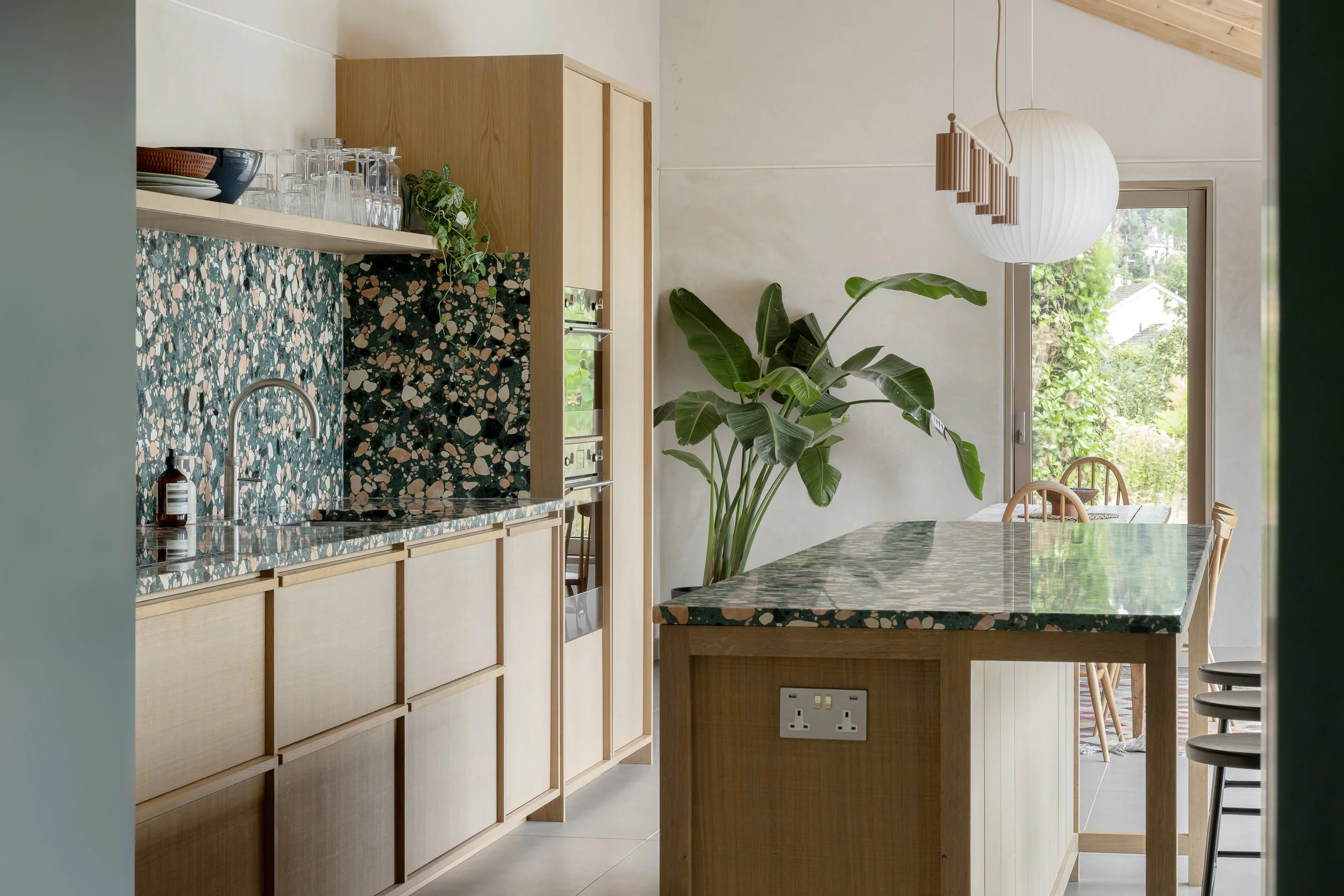 Modern kitchen with terrazzo countertops, light wood cabinets, green leafy plant near large window, and unique pendant lighting.