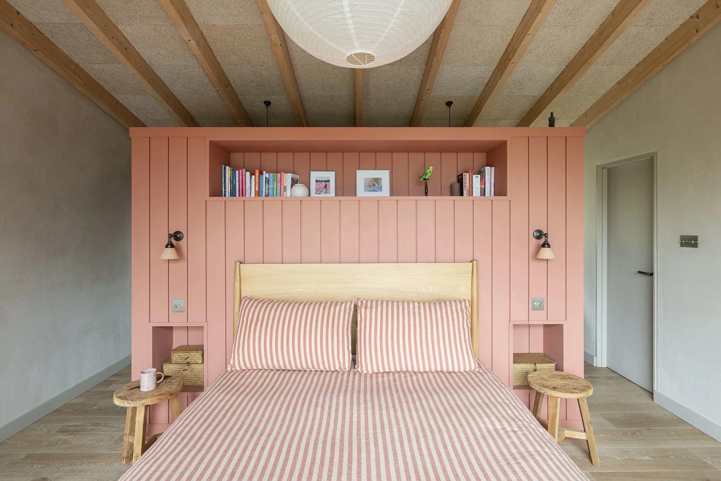 A bedroom with a pink headboard wall, a wooden bed with pink and white striped bedding, two small wooden stools on each side, built-in shelves with books and decorations, wall-mounted lamps, and a ceiling paper lantern.