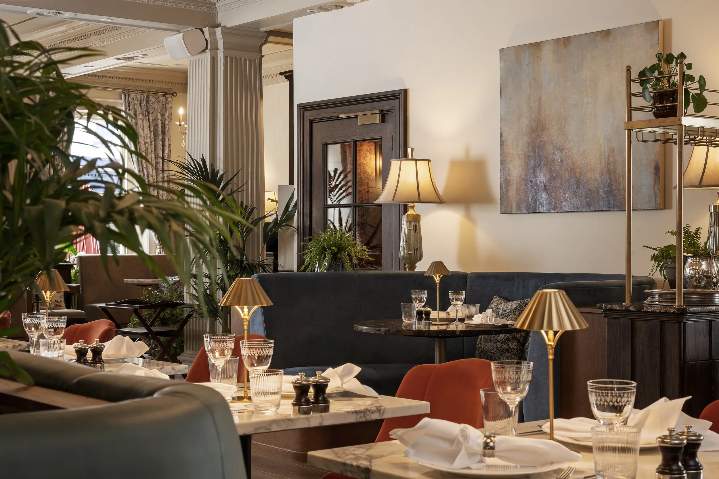 Interior of an elegant restaurant with tables set with glassware, napkins, and small lamps. The decor includes plush seating, potted plants, and artwork on cream-colored walls.
