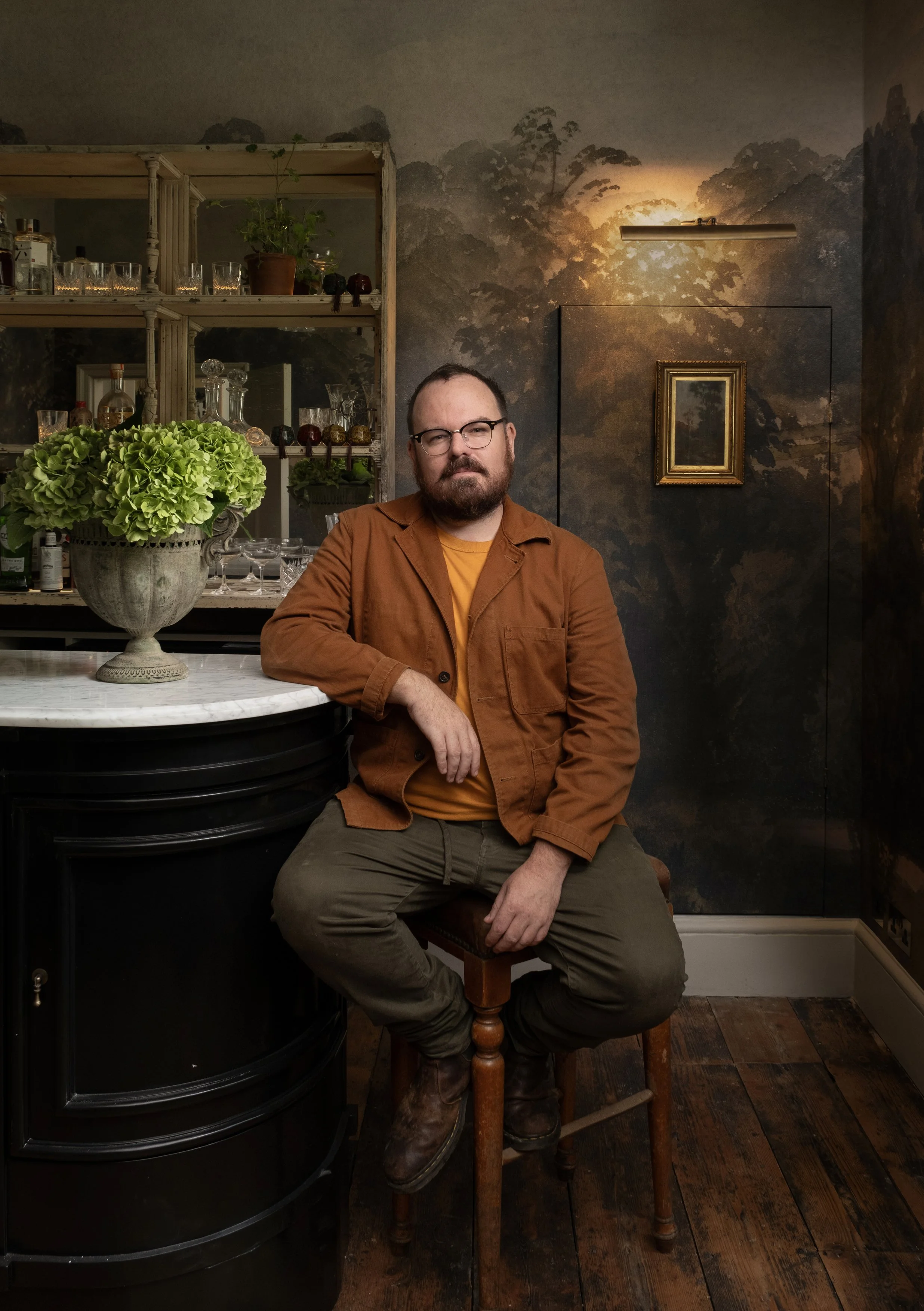 A man with glasses and a beard sitting on a wooden stool in a dimly lit room with dark, forest-themed wallpaper, a bar with glassware, and a large potted green hydrangea on a marble-topped counter.