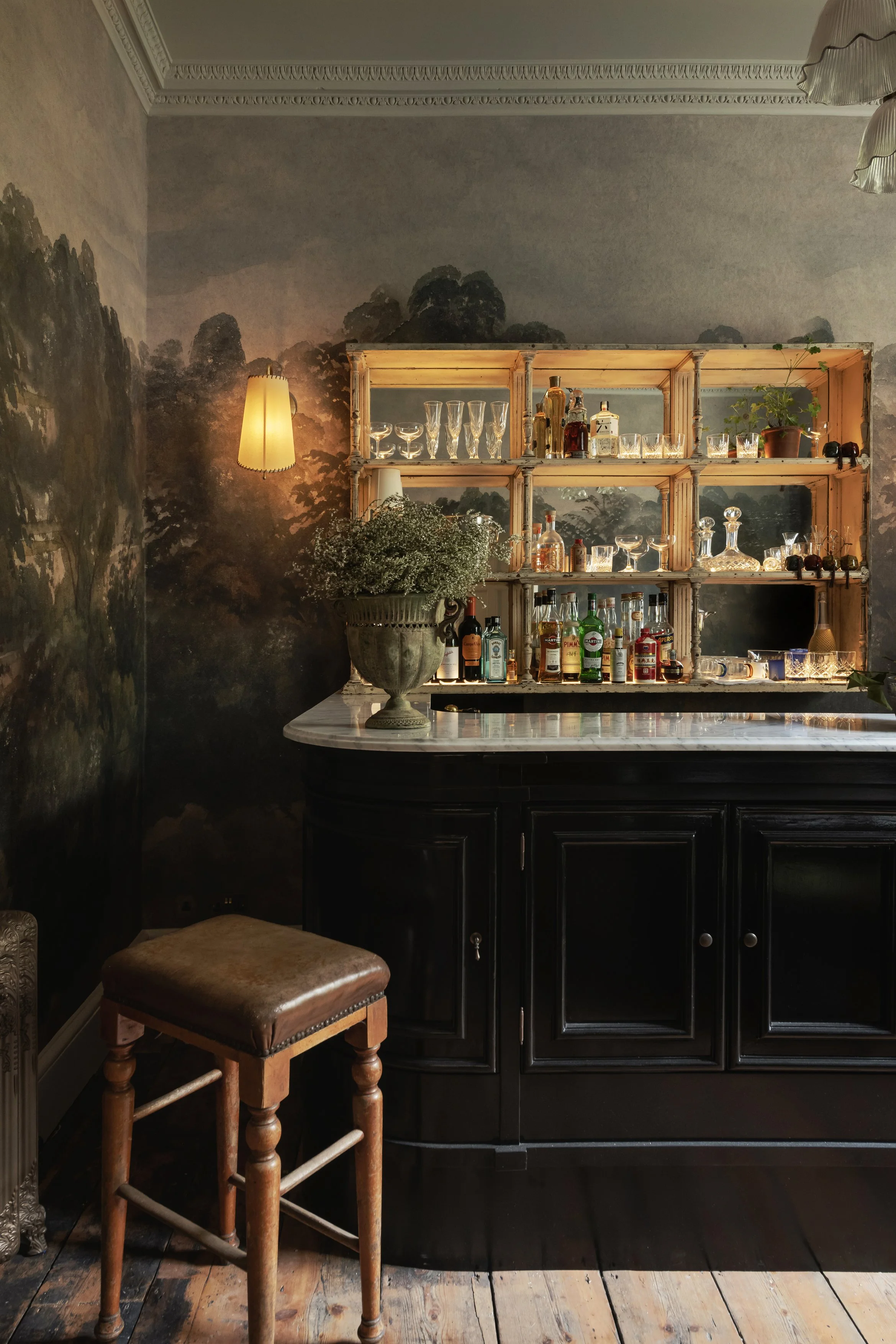 A vintage-style bar with a black cabinet, a white marble countertop, and a wooden shelving unit displaying various glassware and bottles. A lamp with a yellow lampshade and a potted plant are on the bar. The wall behind has a dark, scenic mural of tr