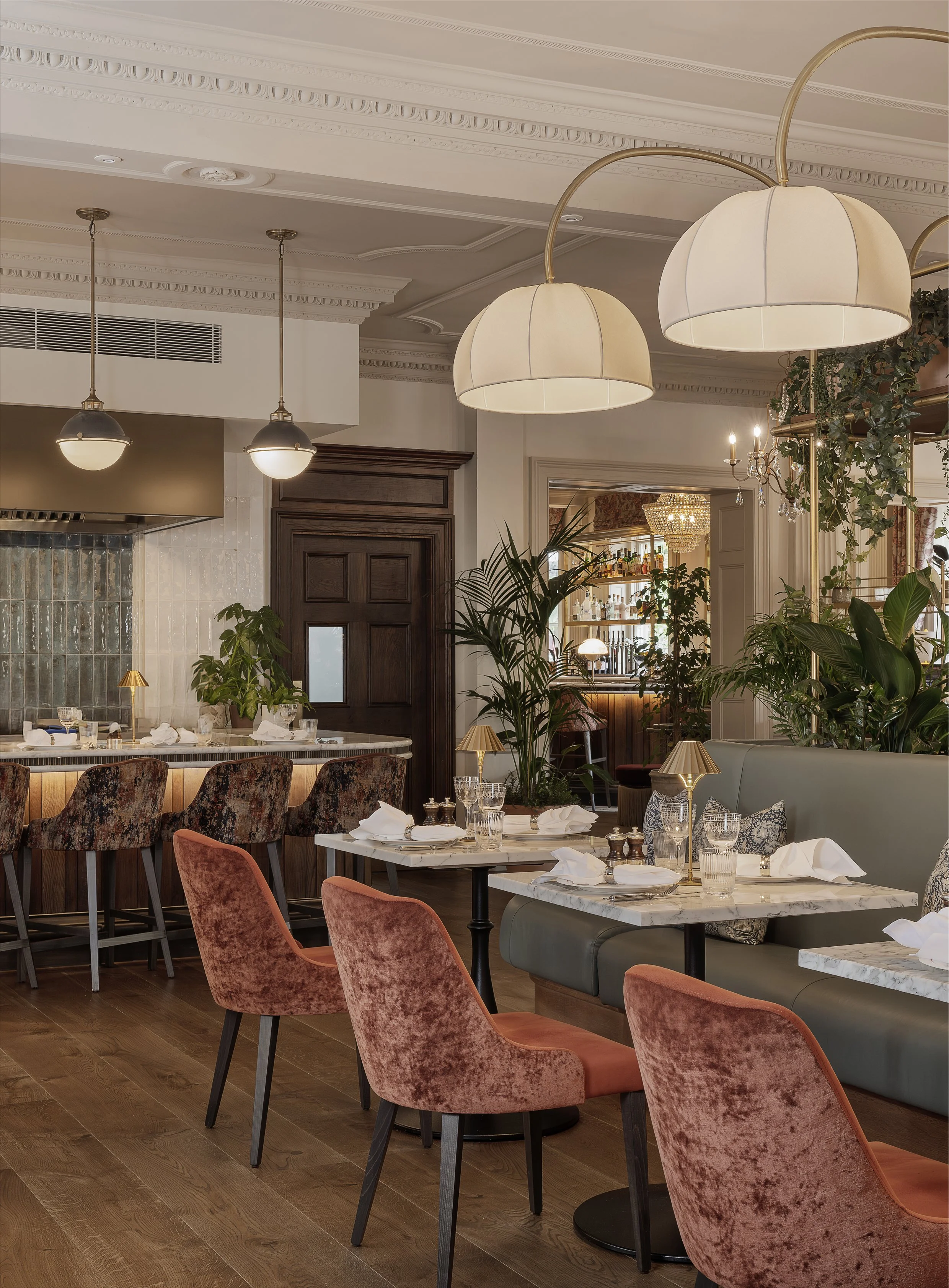 Interior of an upscale restaurant with pink velvet chairs, white marble tables set with white napkins, glassware, and small lamps. Green plants and elegant lighting fixtures are visible, with a bar area in the background.