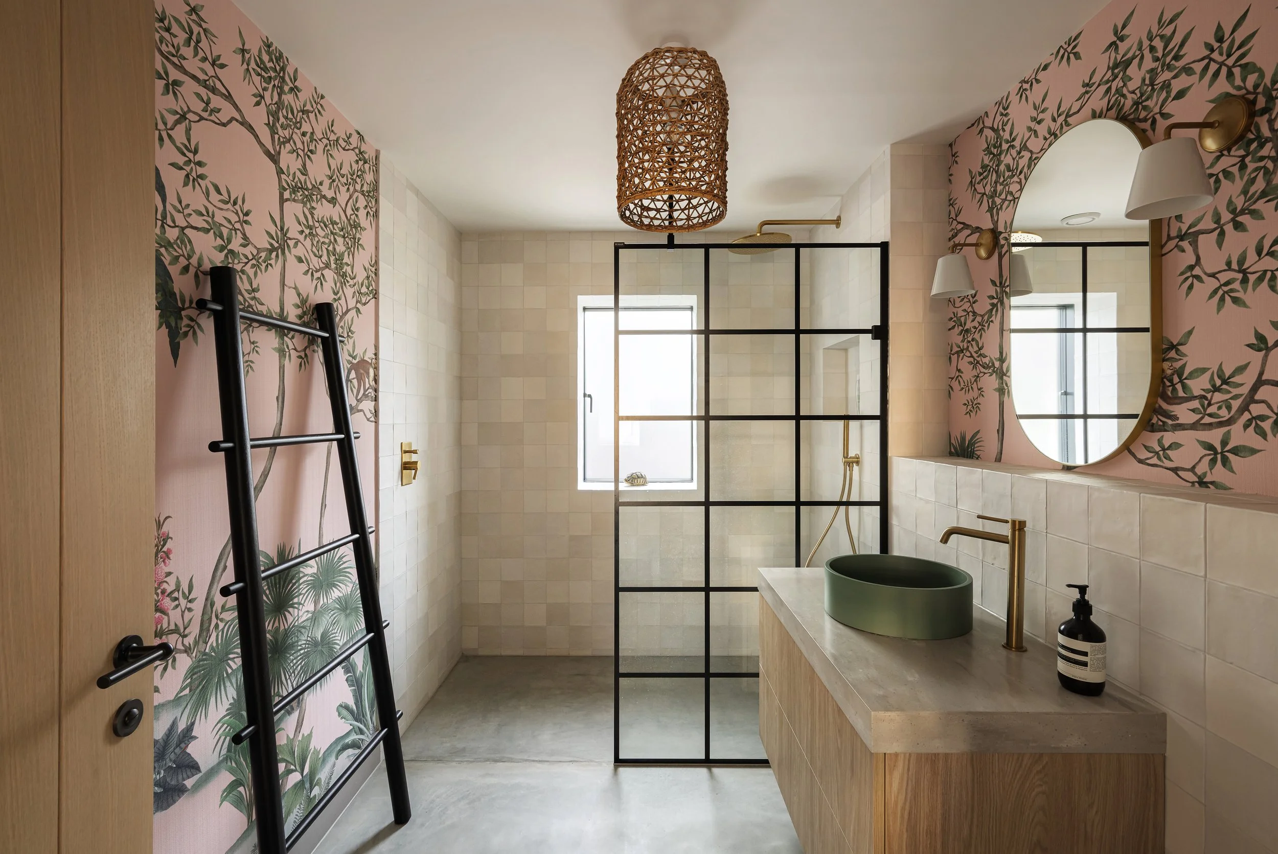 Modern bathroom with pink floral wallpaper, round mirror, black and gold fixtures, green countertop vessel sink, black ladder towel rack, and a glass shower enclosure with black grid framing.