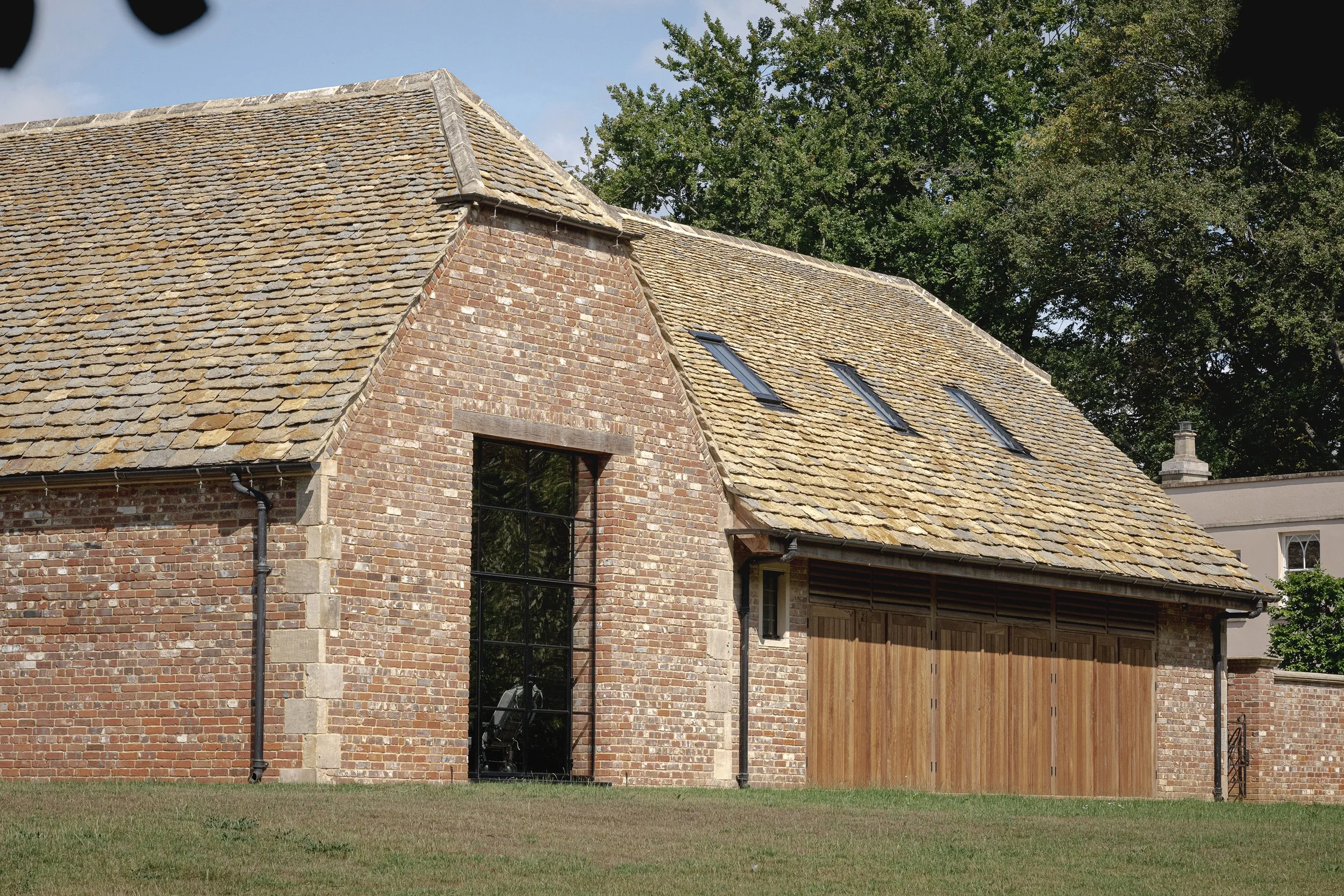 A brick house with a rustic, sloped tile roof, large glass window, and wooden garage door. The roof has three skylights, and there is greenery and trees in the background.