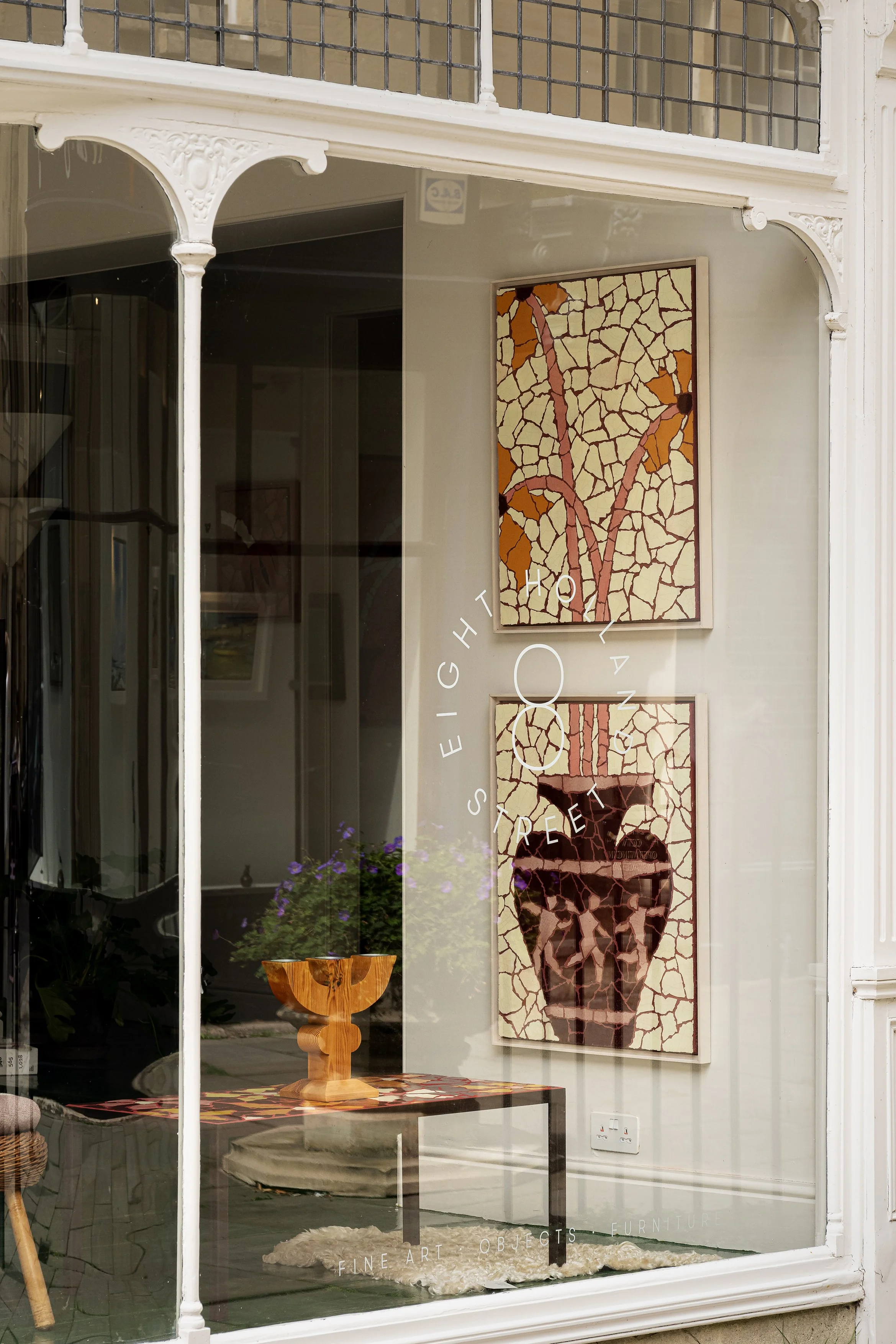 A store window display with artwork and furniture, including two mosaic-style paintings of a vase and leaves, a wooden vase on a glass table, and purple flowers in the background.