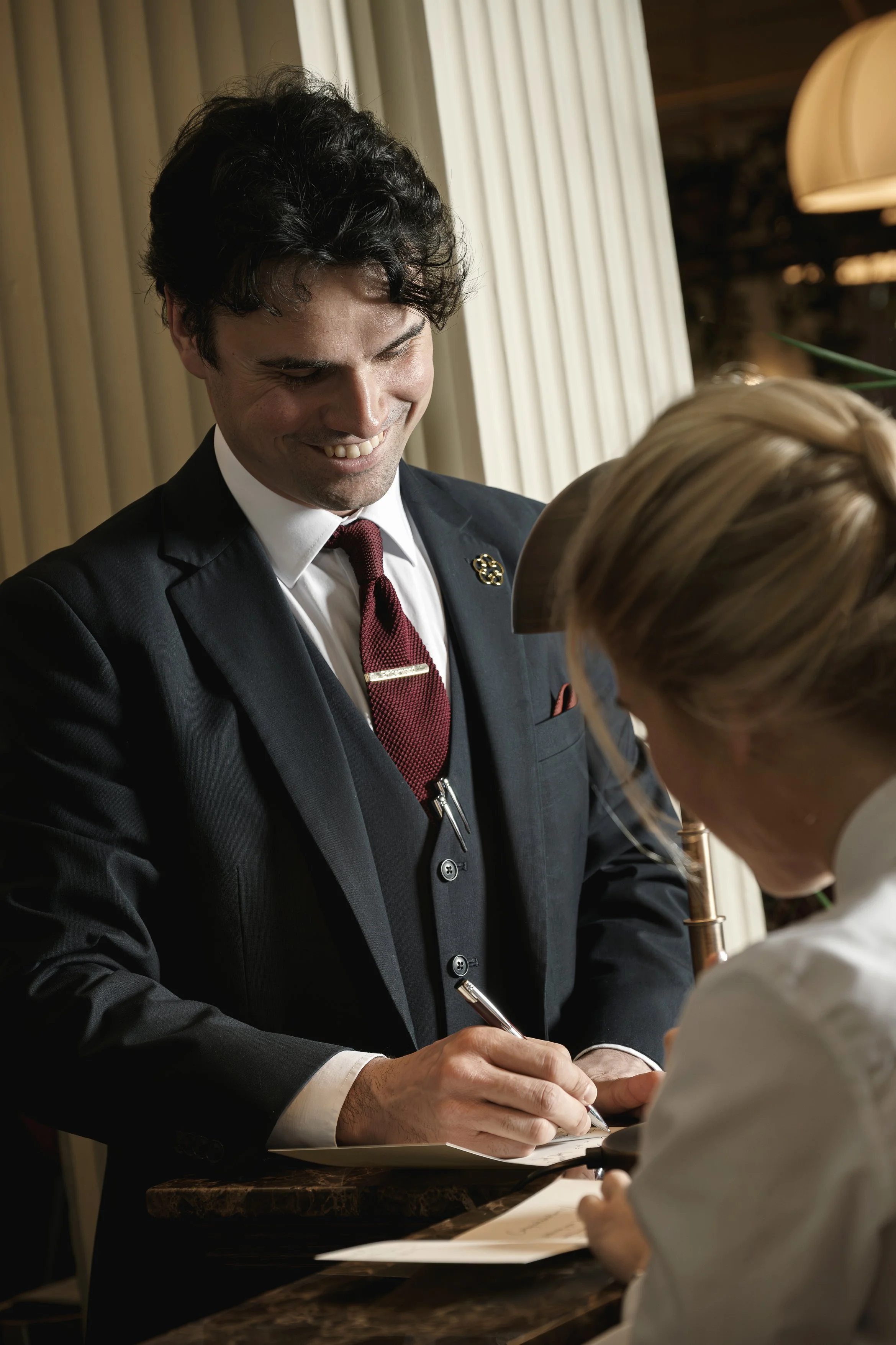 Man in a suit smiling as he signs a document for a blonde woman
