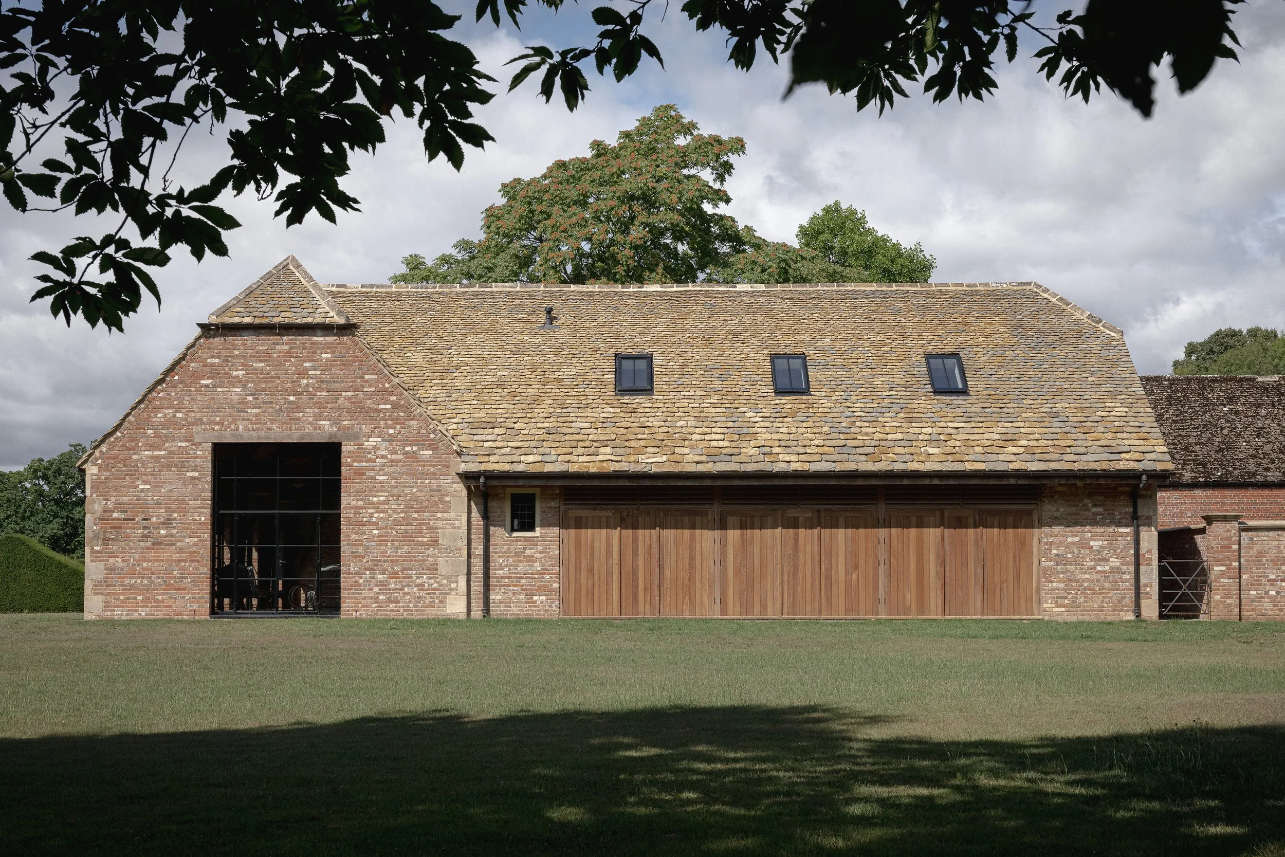A large brick barn with a tiled roof, featuring two small windows and a large wooden door, set against a cloudy sky and surrounded by green trees and grass.