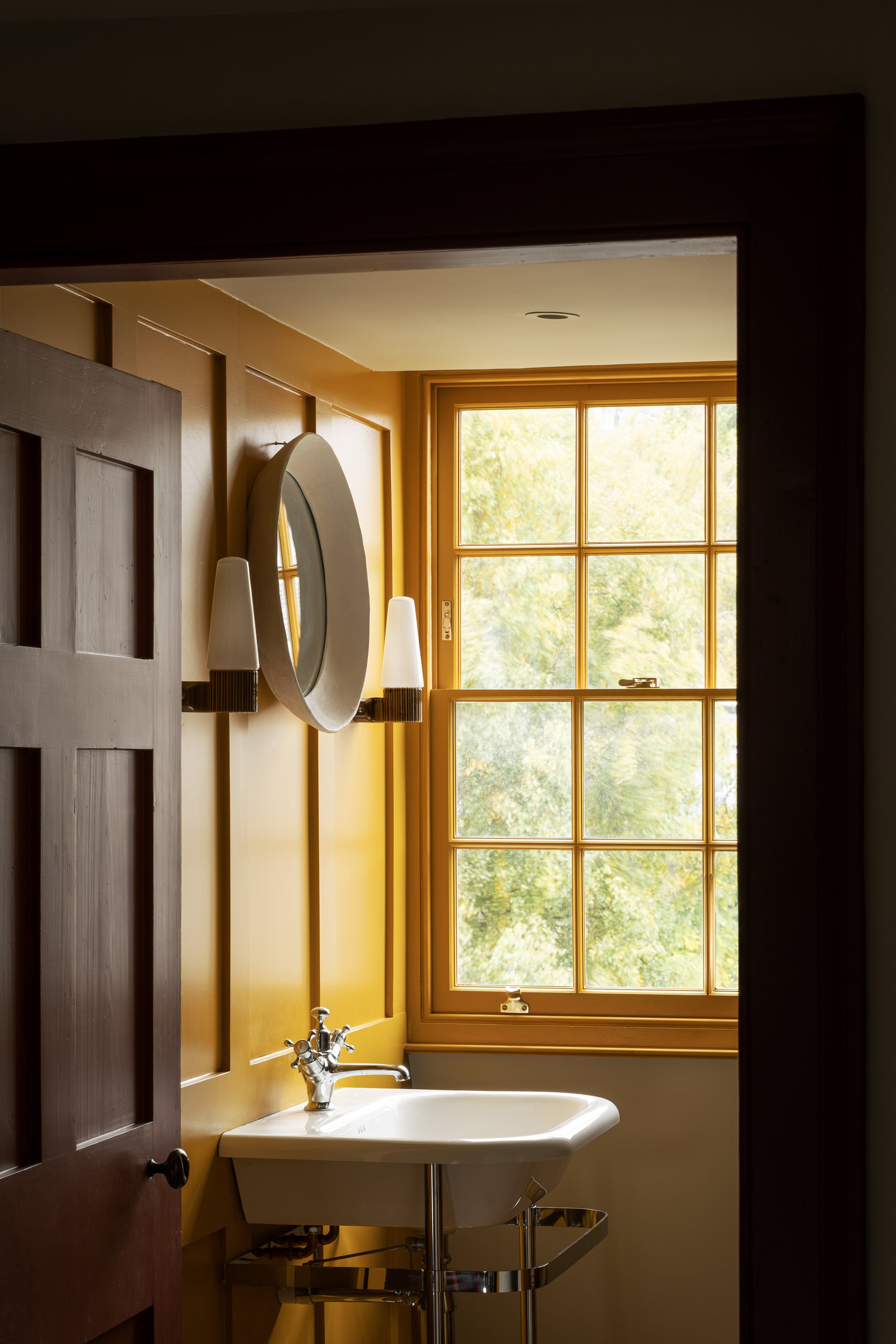 Interior view of a small bathroom with a yellow wall, a large window with a yellow frame, a white sink, and a mirror with two wall sconces.