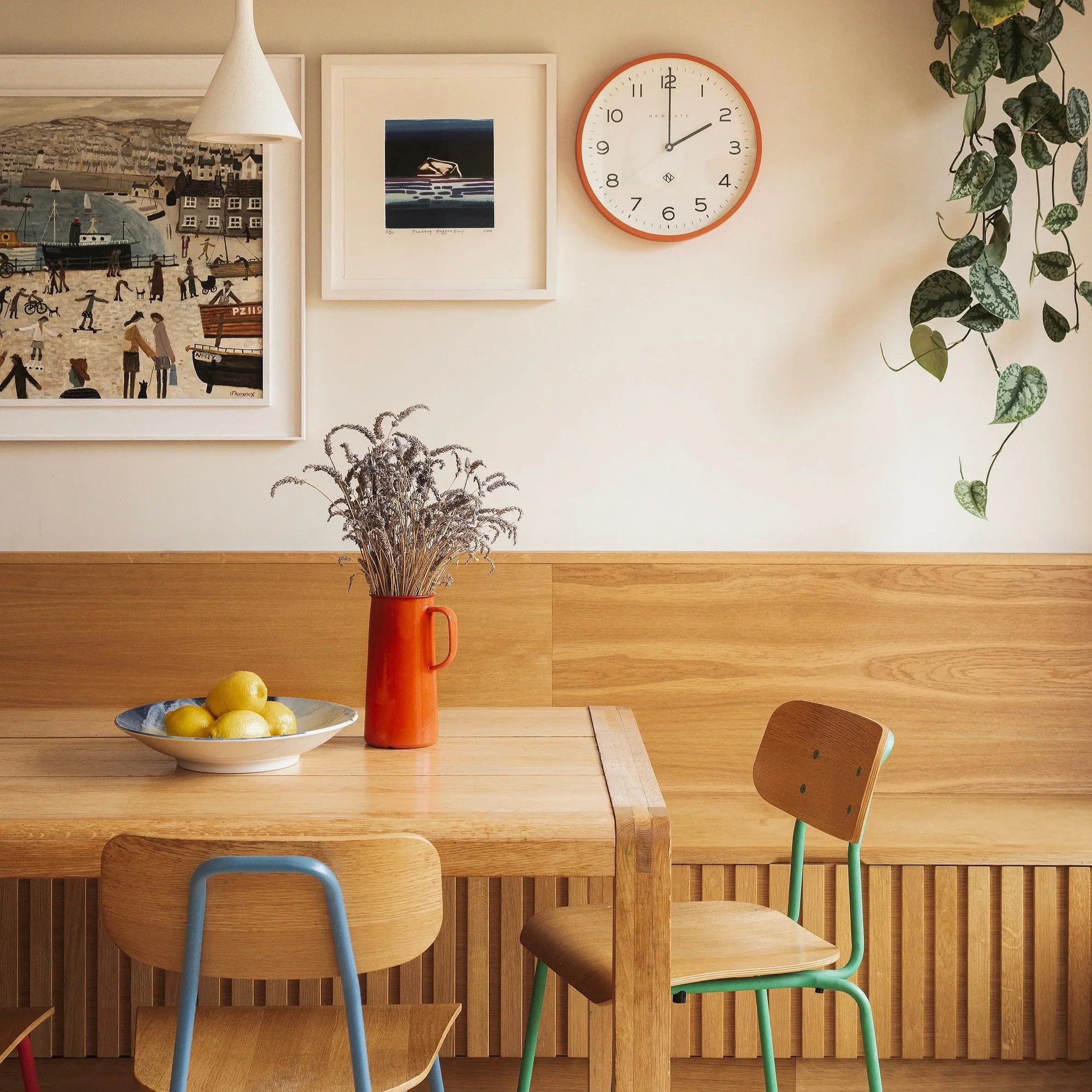 A dining area with a wooden table, a plant in a red vase, a bowl of lemons, and colorful chairs. The background features a wall with framed artwork, a clock, and hanging plants.