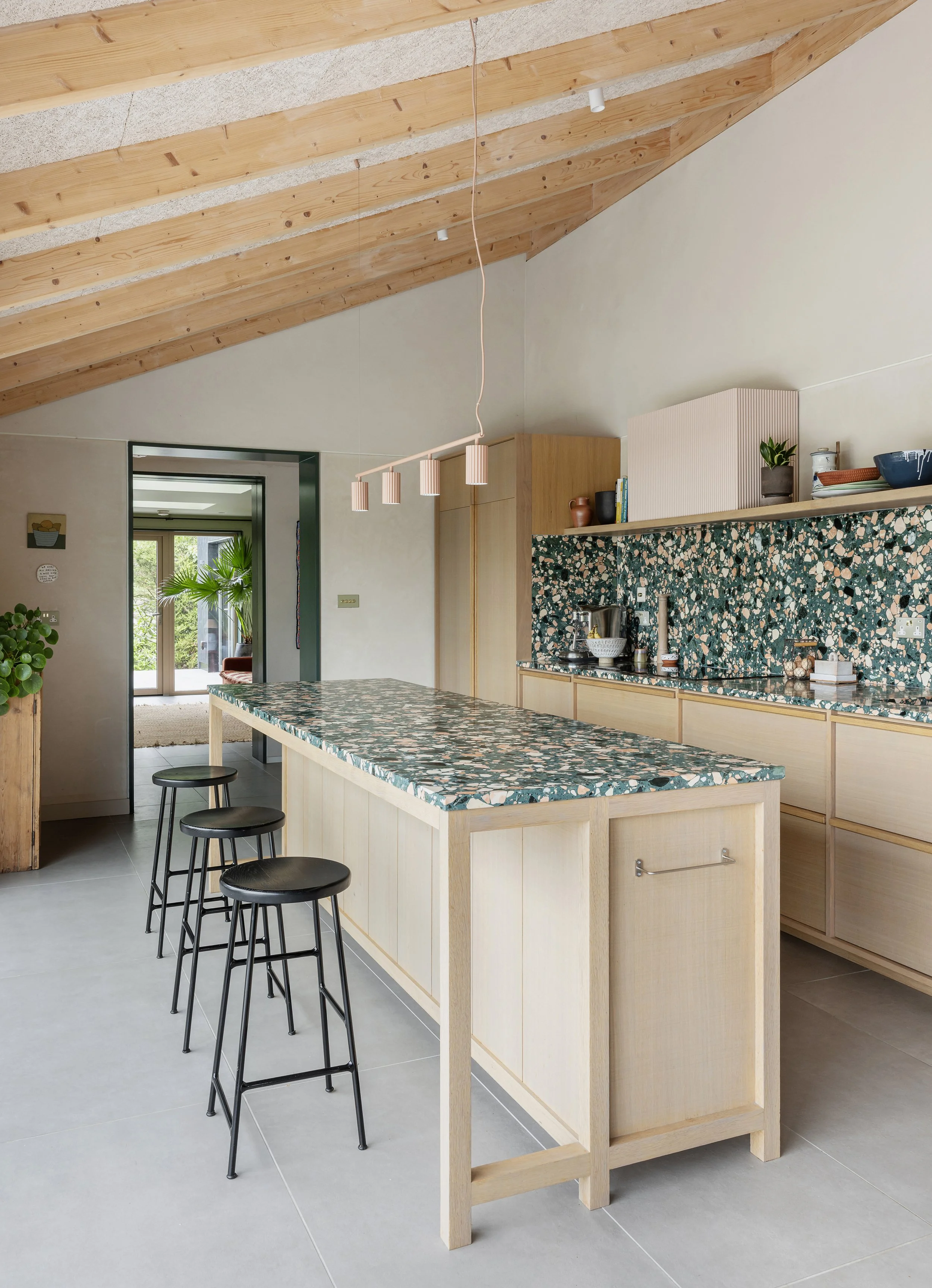 Modern kitchen with a terrazzo island, wooden cabinetry, and a terrazzo backsplash. There are three black stools at the island and a sliding glass door leading outside.