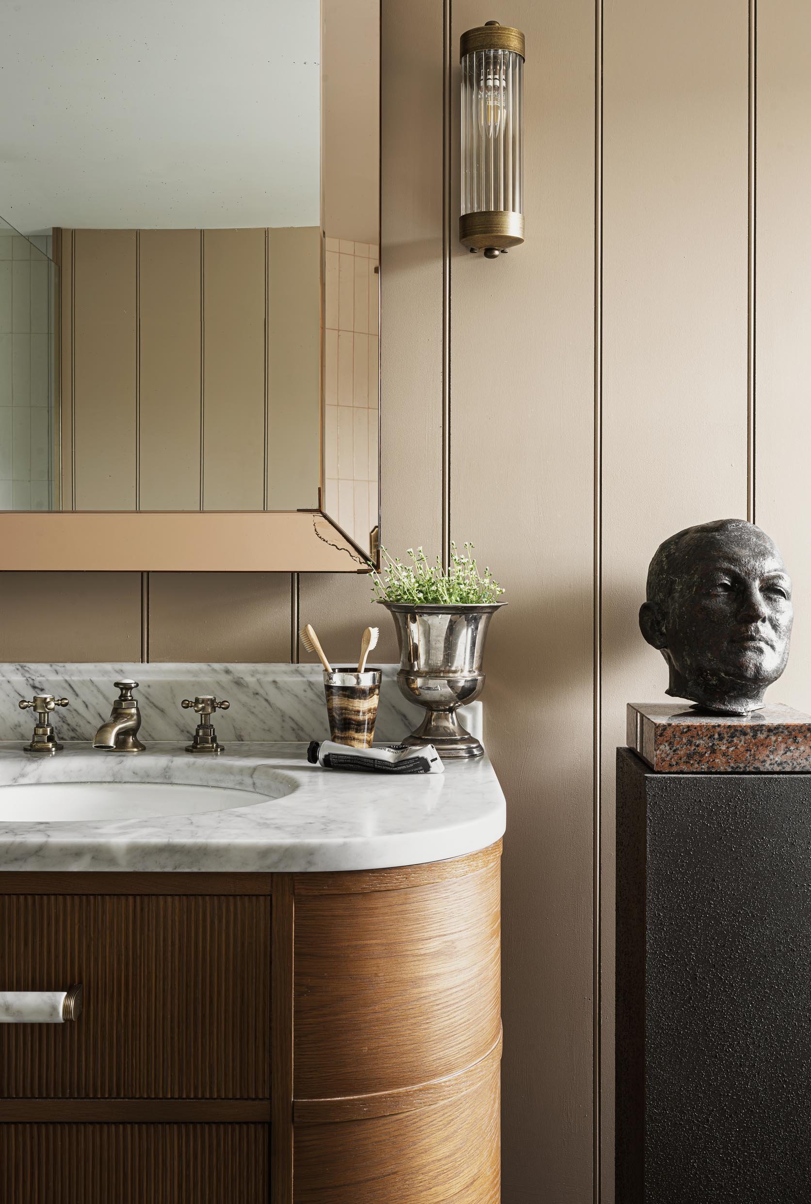 A bathroom vanity with a marble countertop, brass faucet, a small plant in a silver vase, toothbrushes in a striped holder, and a tube of toothpaste. A large mirror on the wall shows part of the room. On the right, there is a black sculpture of a hum