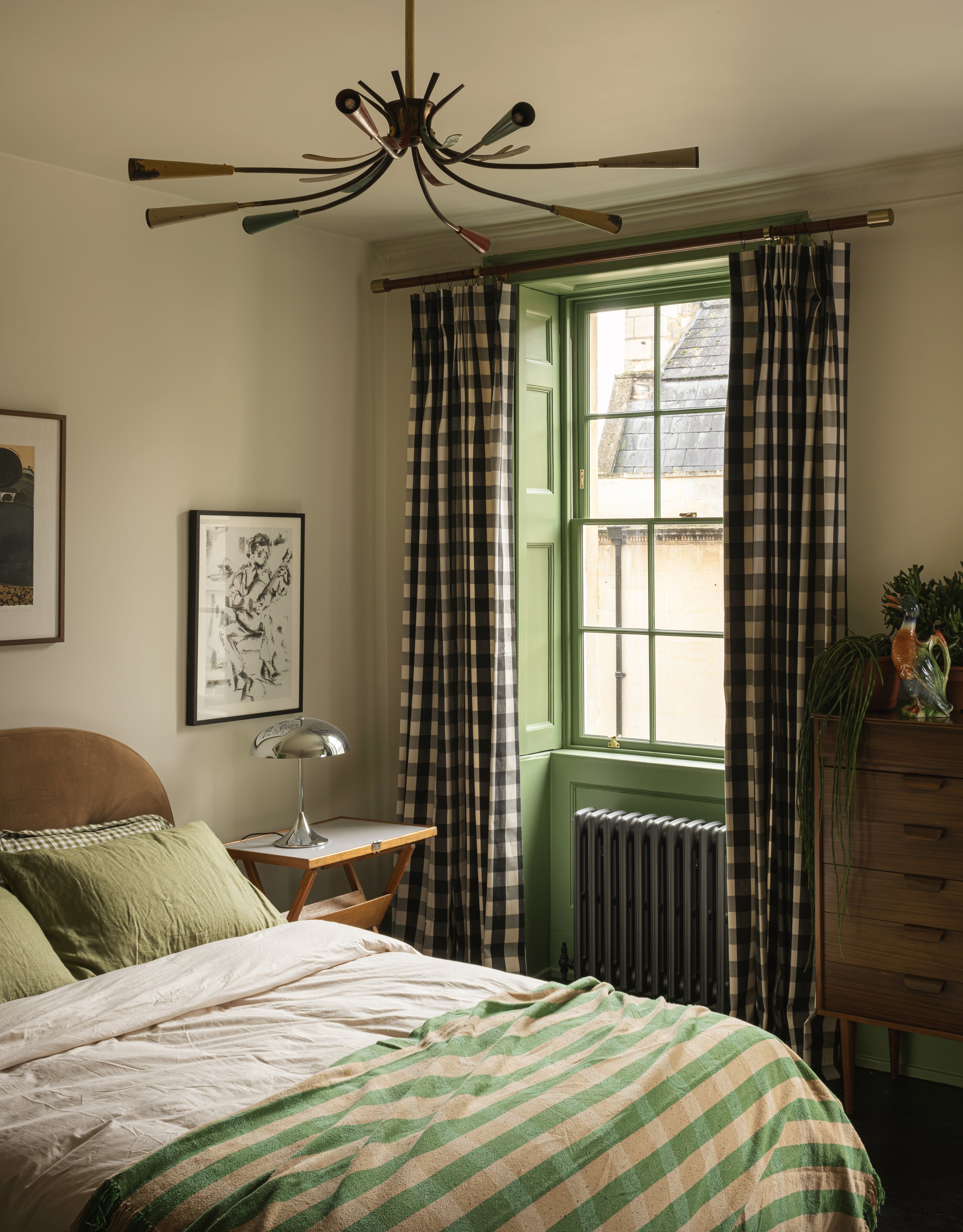 A cozy bedroom with a bed that has green pillows and a striped blanket, an old-fashioned lamp on a wooden nightstand, a window with green shutters and checkered curtains, and a wooden dresser with plants and decorative items.