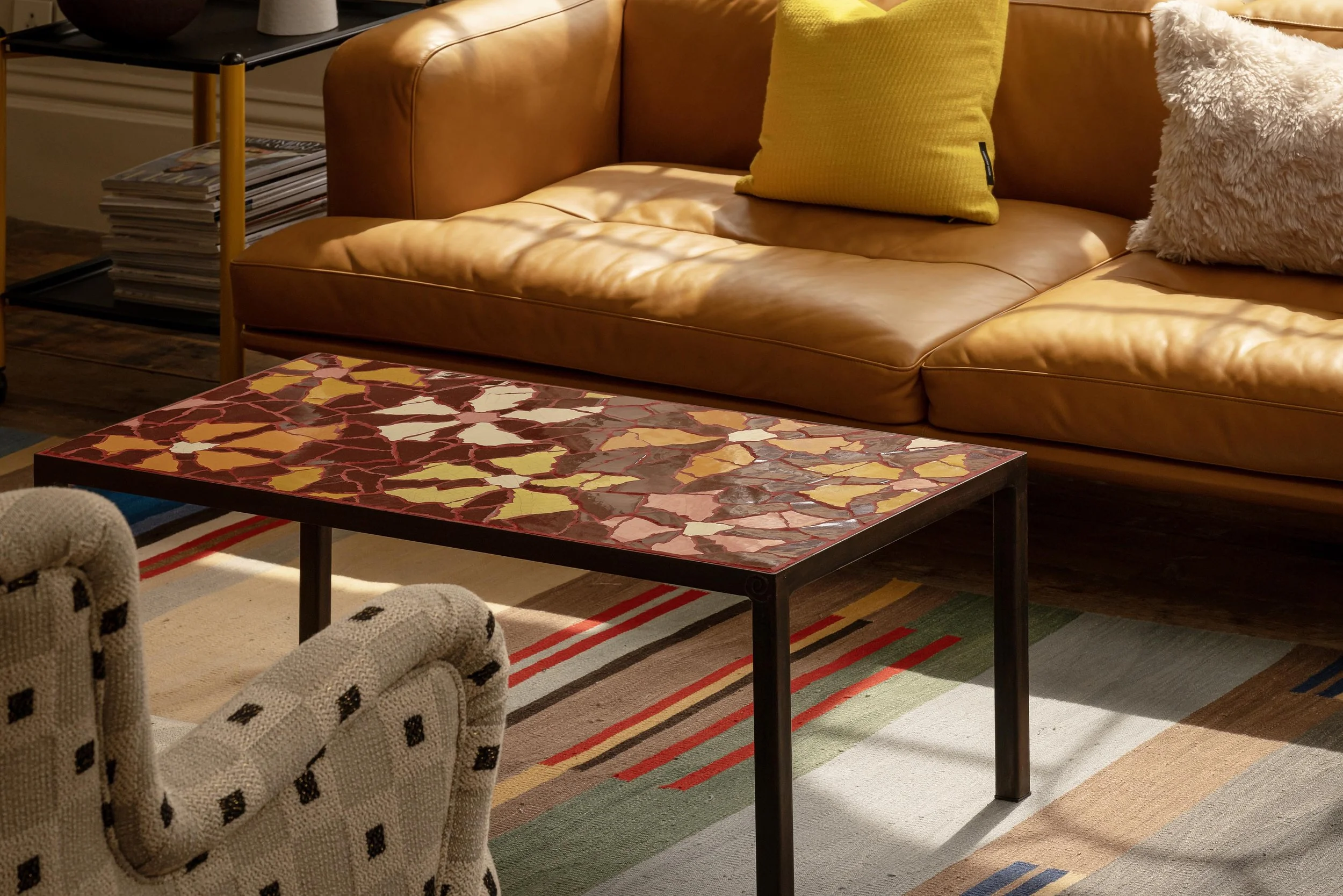 Living room with a tan leather sofa, yellow and beige textured cushions, a colorful patchwork coffee table, a patterned area rug, and a side table with magazines.