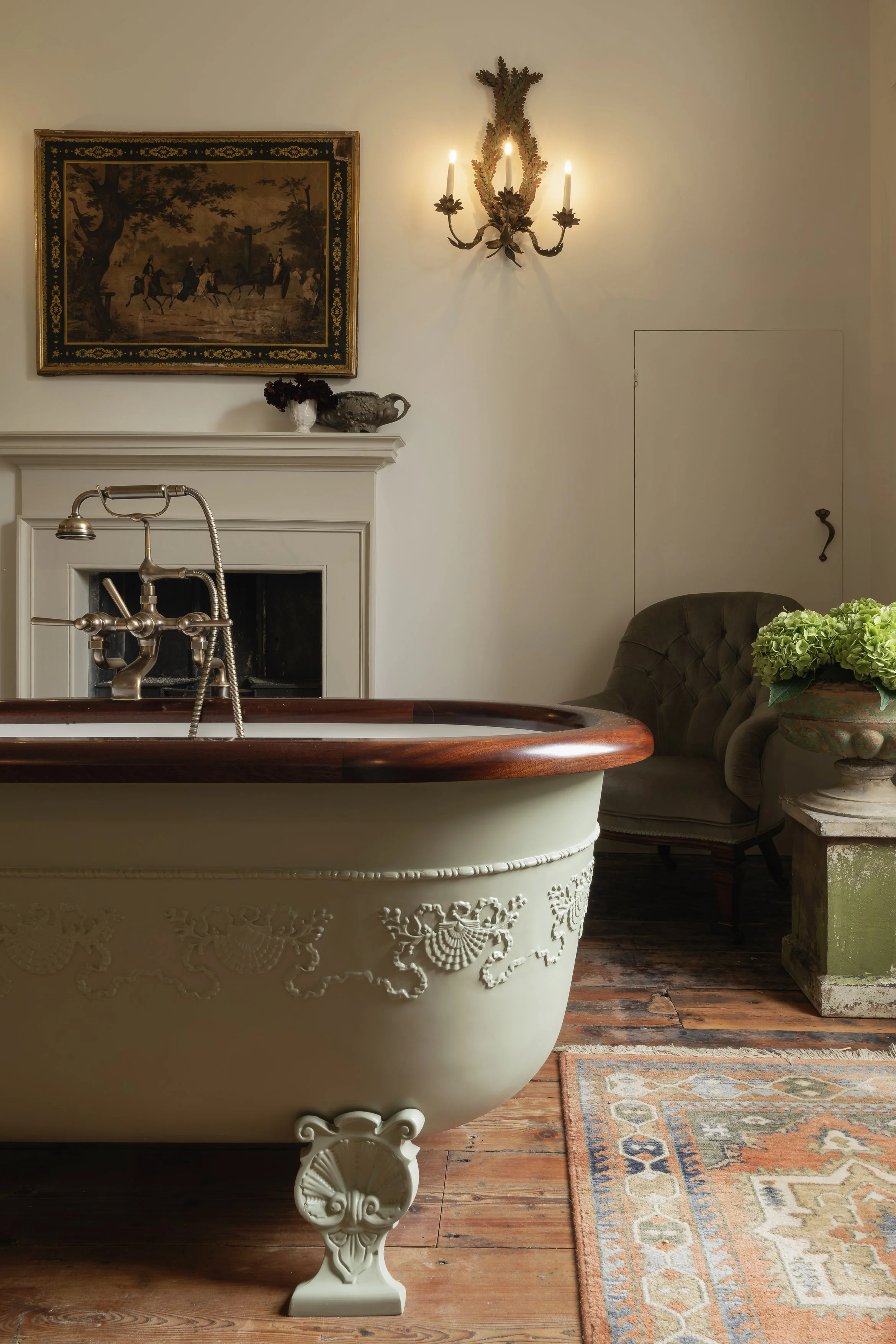 A vintage bathroom with a clawfoot bathtub, a dark green upholstered armchair, a small side table with a potted plant, a painting of a hunting scene, a decorated wall sconce, and a patterned area rug on wooden flooring.