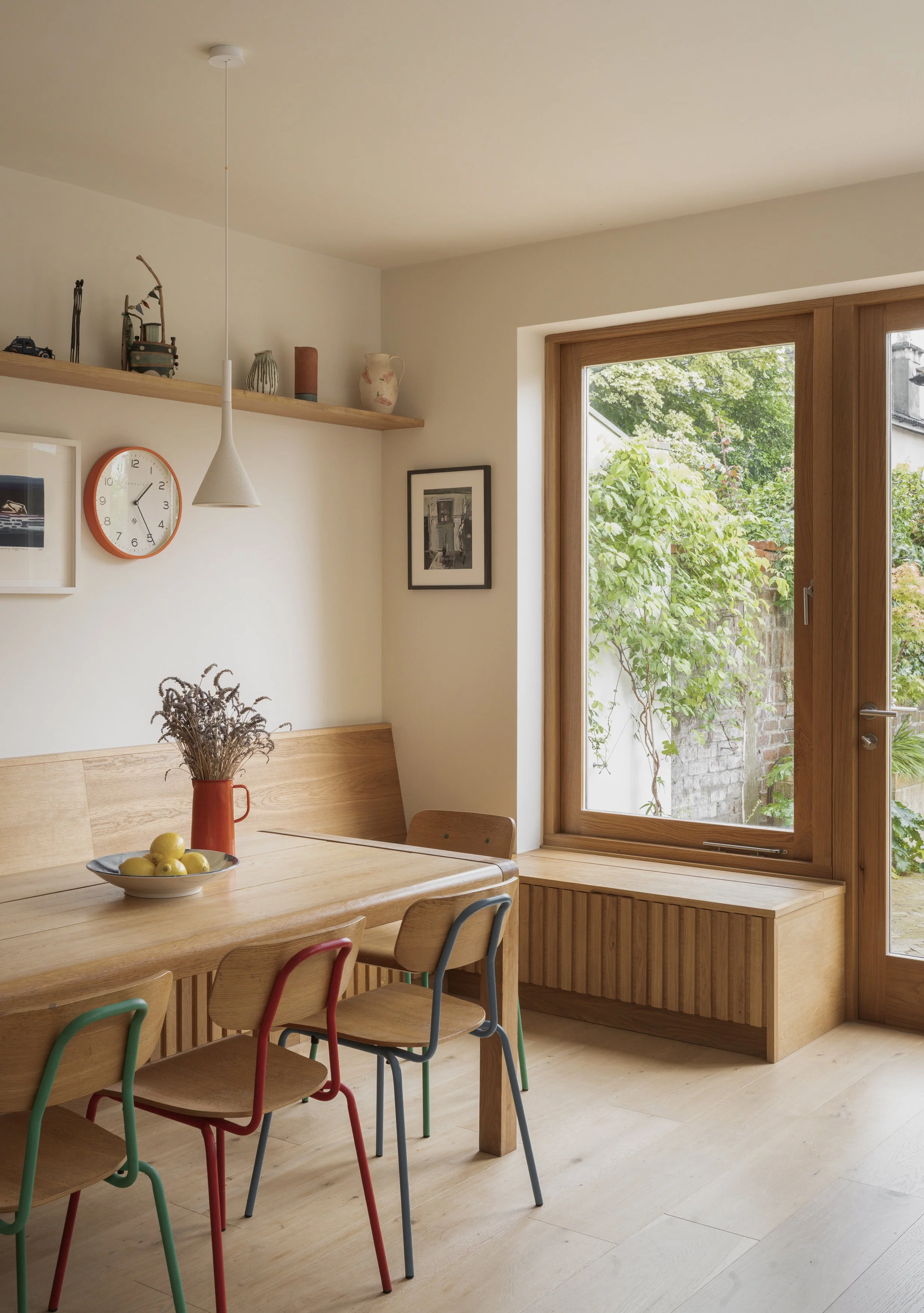 A dining area with a wooden table and colorful chairs, a window with a wooden frame showing greenery outside, a wall shelf with decorative items, and a clock on the wall.
