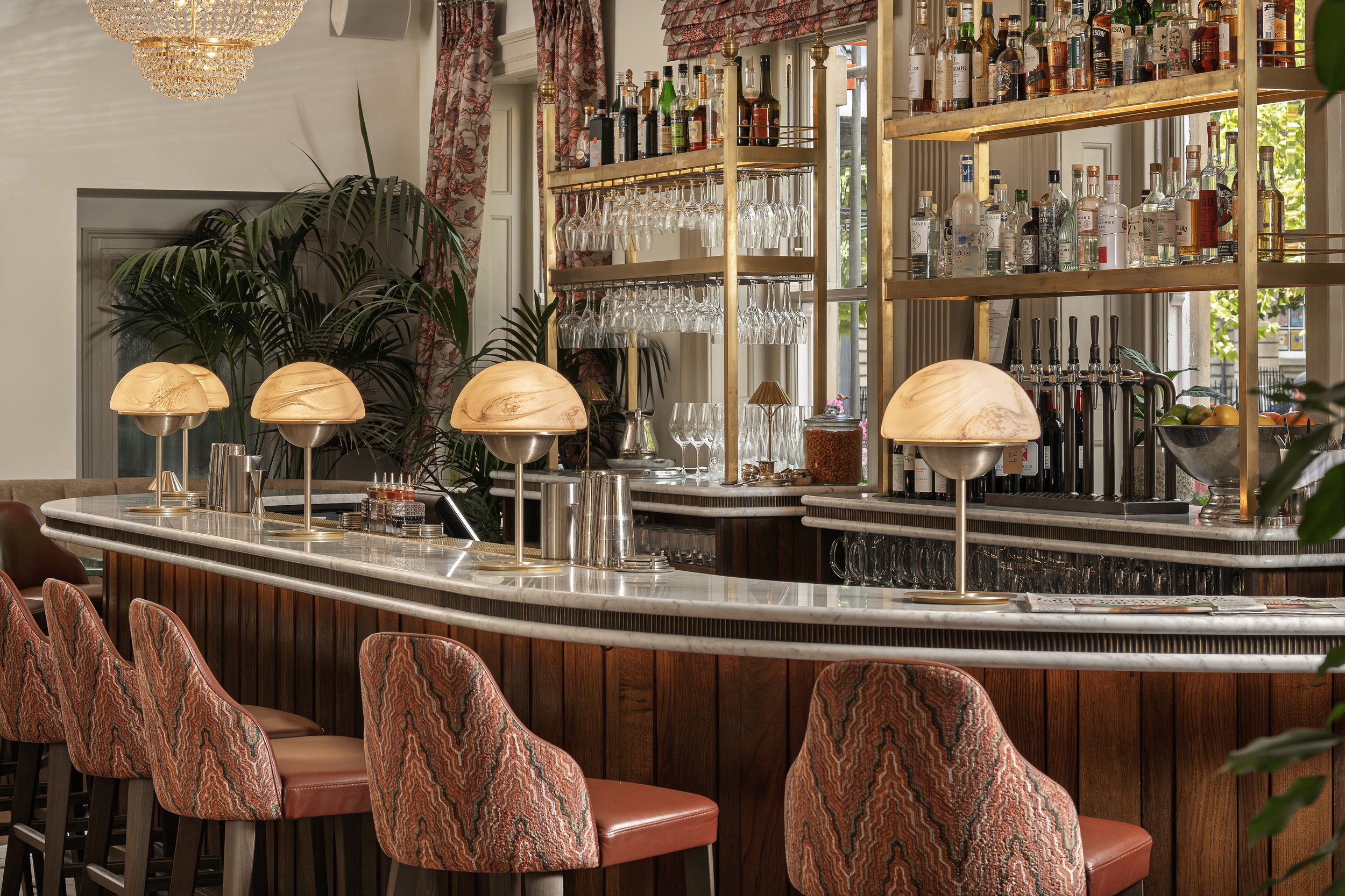 Elegant bar with marble counter, vintage patterned chairs, and glass shelving holding alcohol bottles and glassware, with large windows and lush green plants in the background.