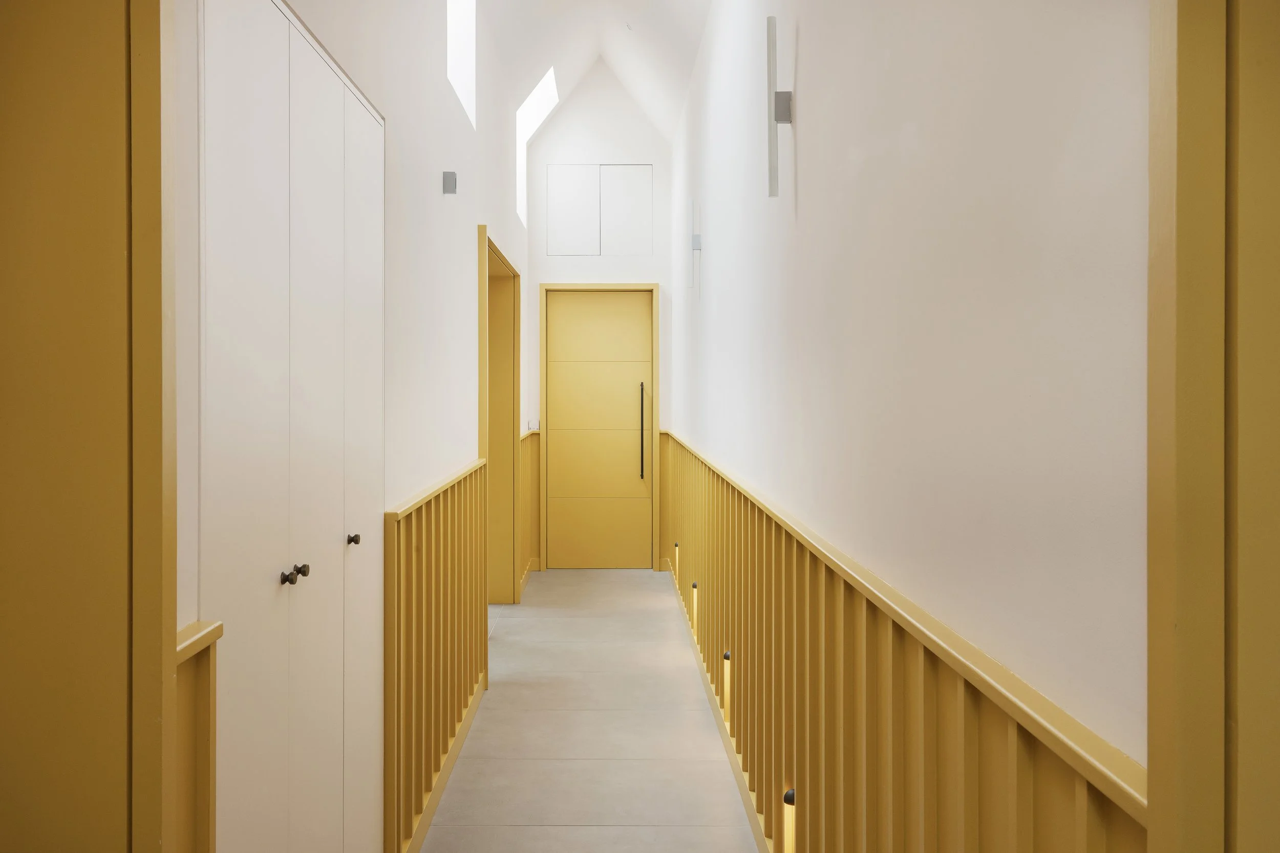 Bright, minimalist hallway with white and yellow walls, a yellow door at the end, and skylights illuminating the space.
