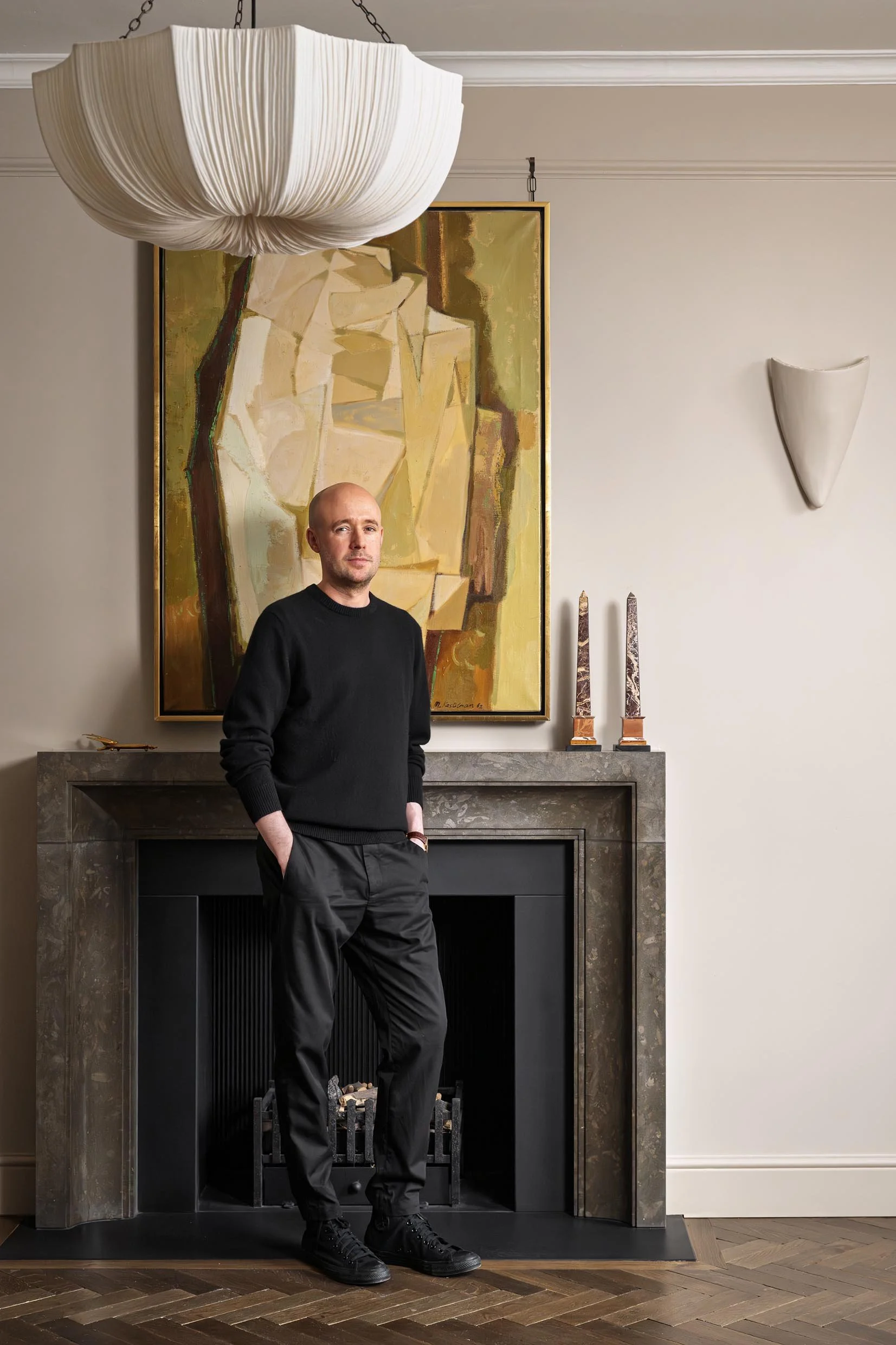 A man with a shaved head wearing a black sweater and black pants standing in front of a modern fireplace with a large abstract artwork hanging above it. The room has beige walls, decorative candles on the mantle, and a large pendant light hanging fro