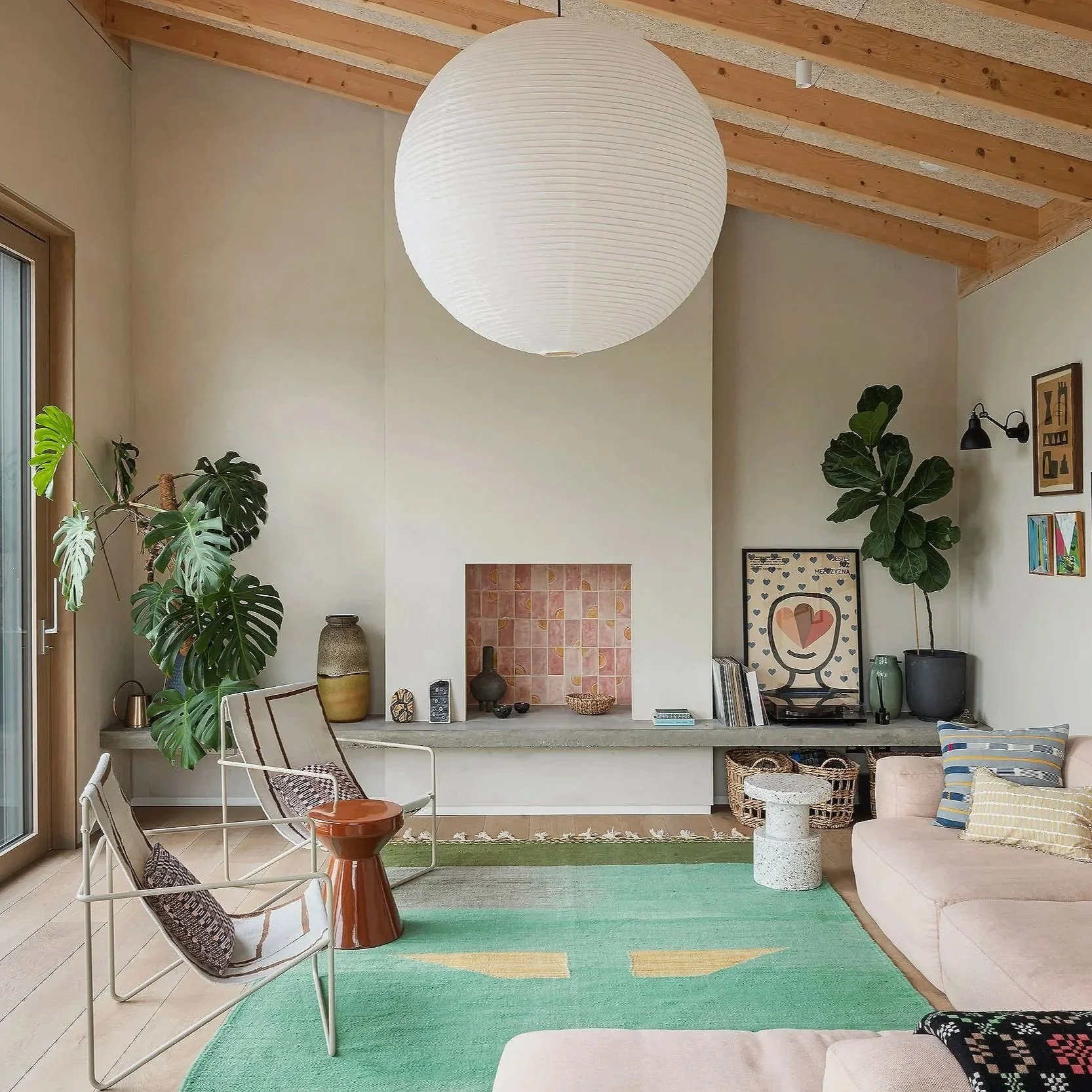 A cozy living room with a high ceiling featuring wooden beams, a large paper lantern pendant light, a fireplace with decorative tiles, indoor plants including a large monstera, and a mix of modern and eclectic furniture, including a pinkish sofa, patterned throw pillows, and a green area rug.