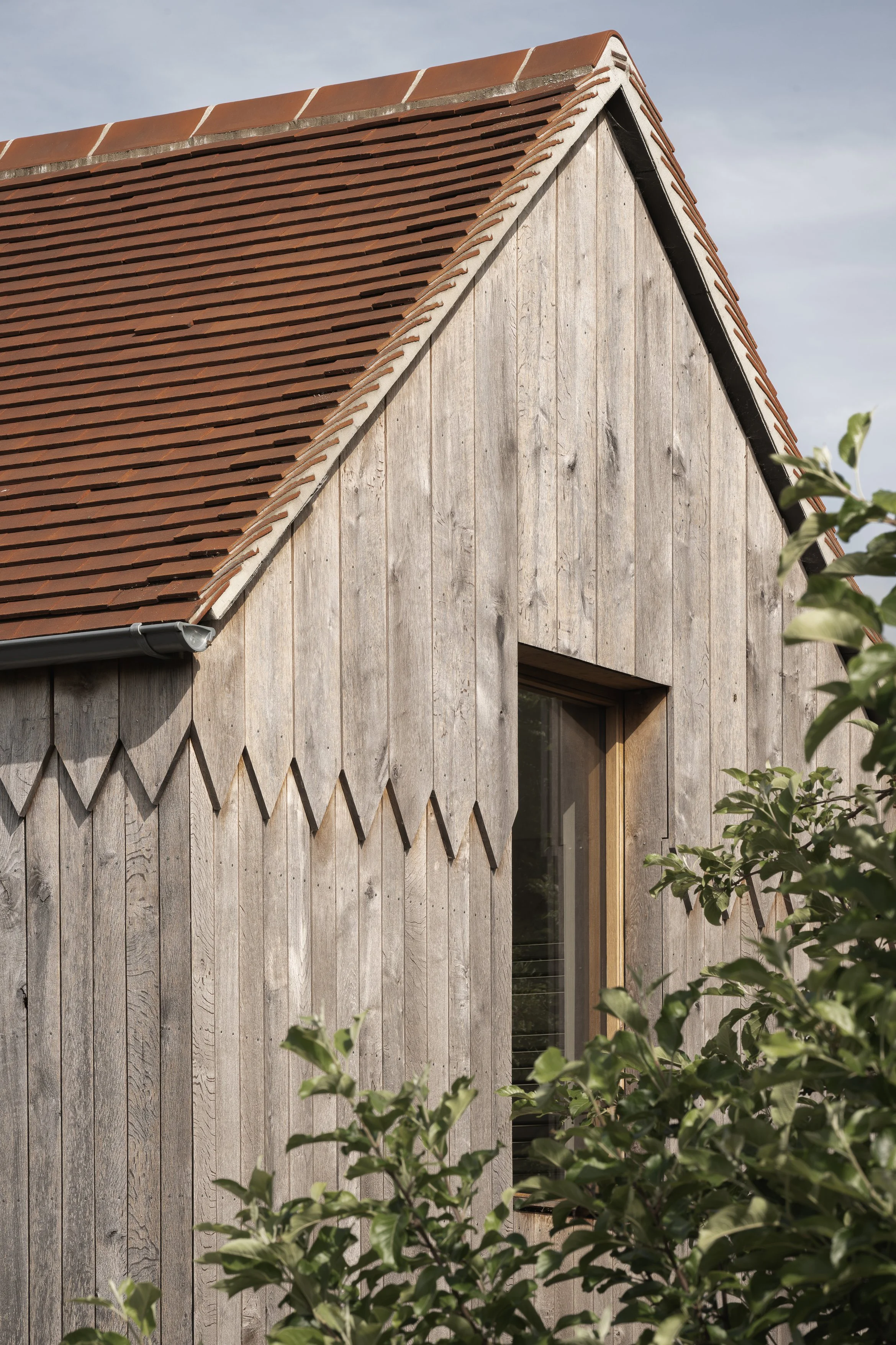 Close-up of a wooden house with vertical planks, a window, and a red-tiled roof. Green leaves in the foreground.