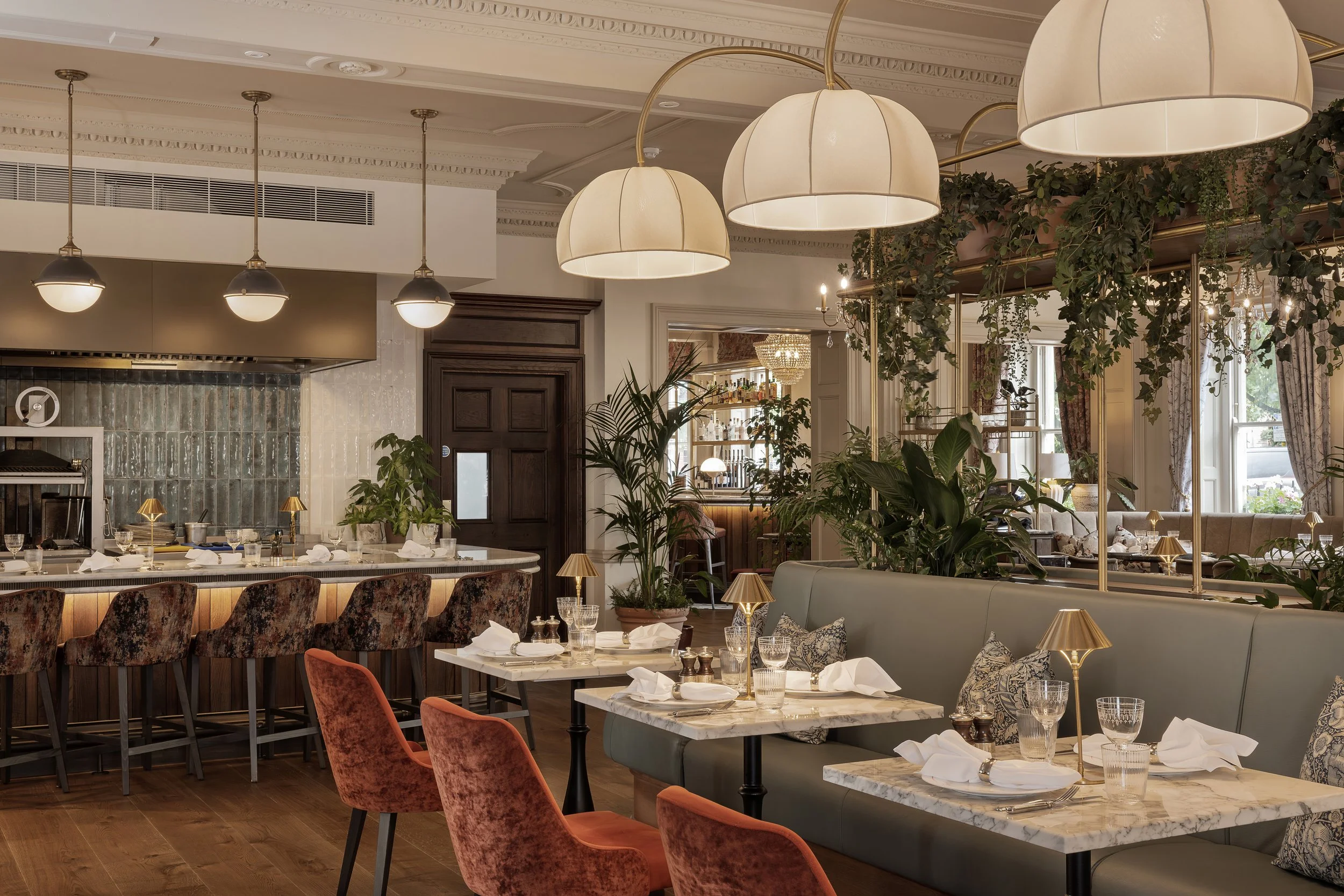 Elegant restaurant interior with hanging lamps, marble-topped tables set with white napkins and glassware, plush pink and blue seating, and green plants.