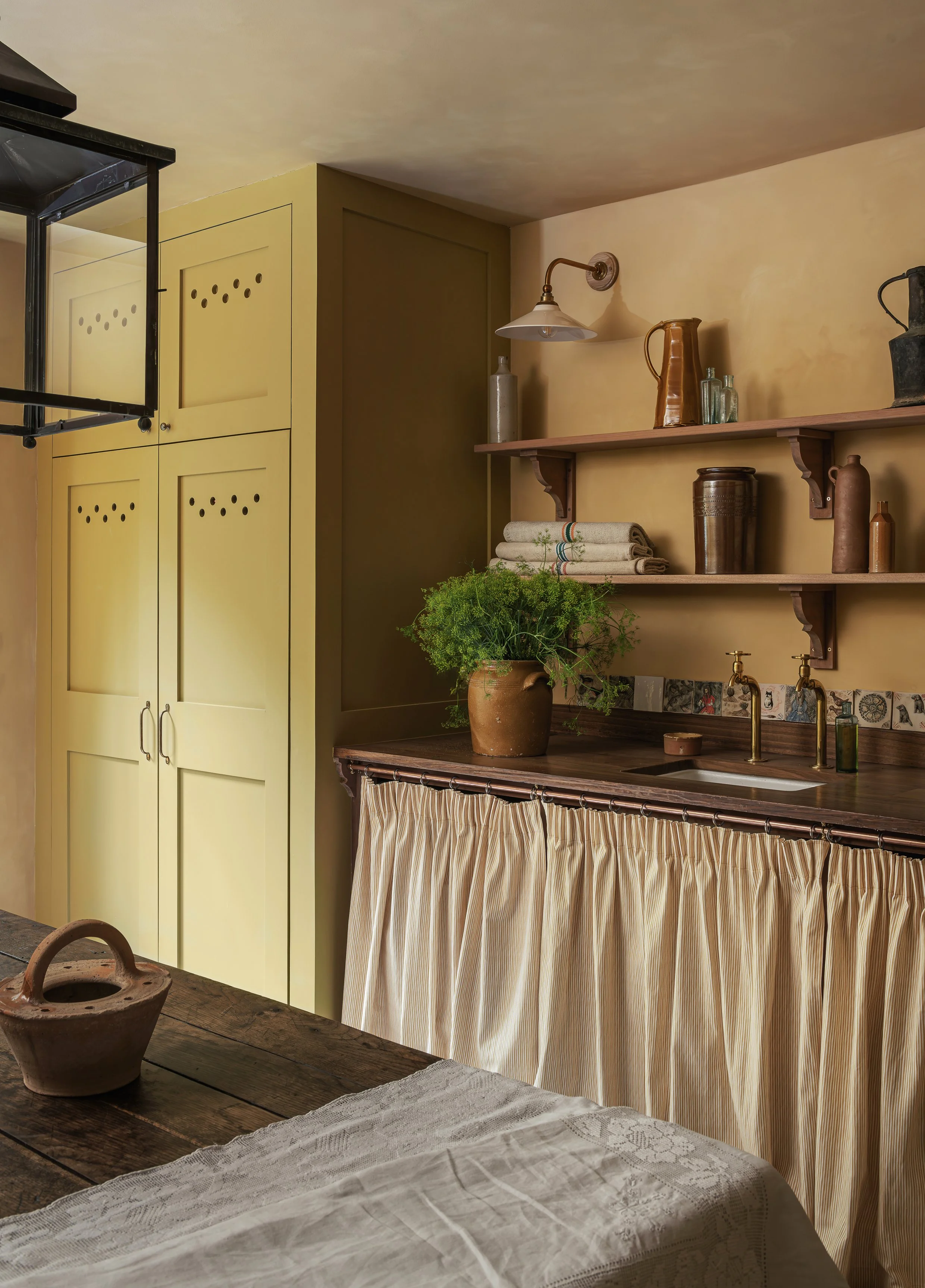 A rustic kitchen corner with yellow cabinets, wooden shelves holding ceramic pitchers and jars, a potted green plant, a farmhouse sink, brass faucet, and a curtain covering the lower part of the counter.