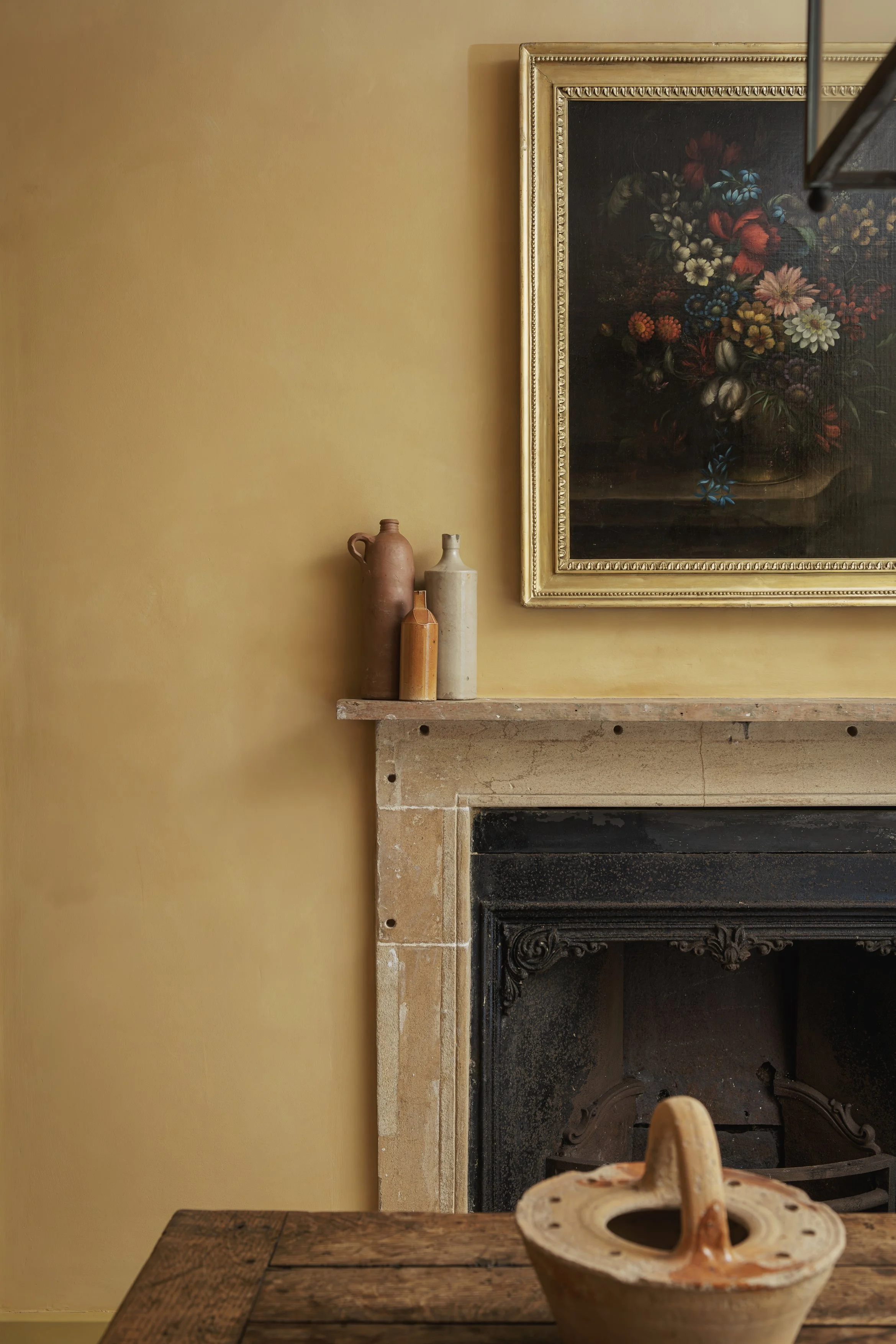 A cozy interior with a yellow wall, a stone fireplace with ornate black detailing, a wooden mantel with three ceramic vases, a framed floral painting, and a rustic wooden table with a pottery bowl in the foreground.
