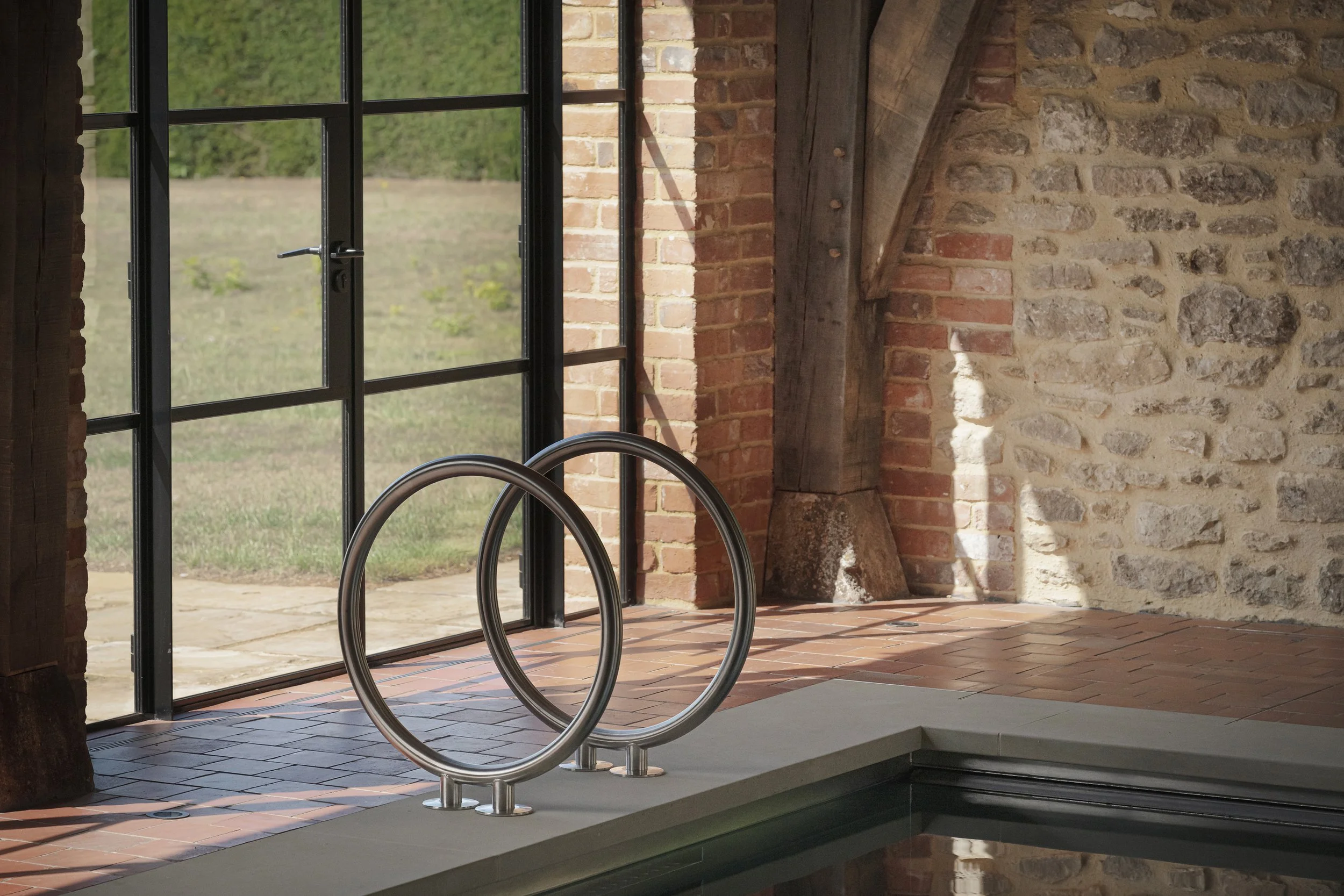 Indoor pool area with metal handrails, brick and stone walls, large glass door showing outdoor grass, and wooden beams.