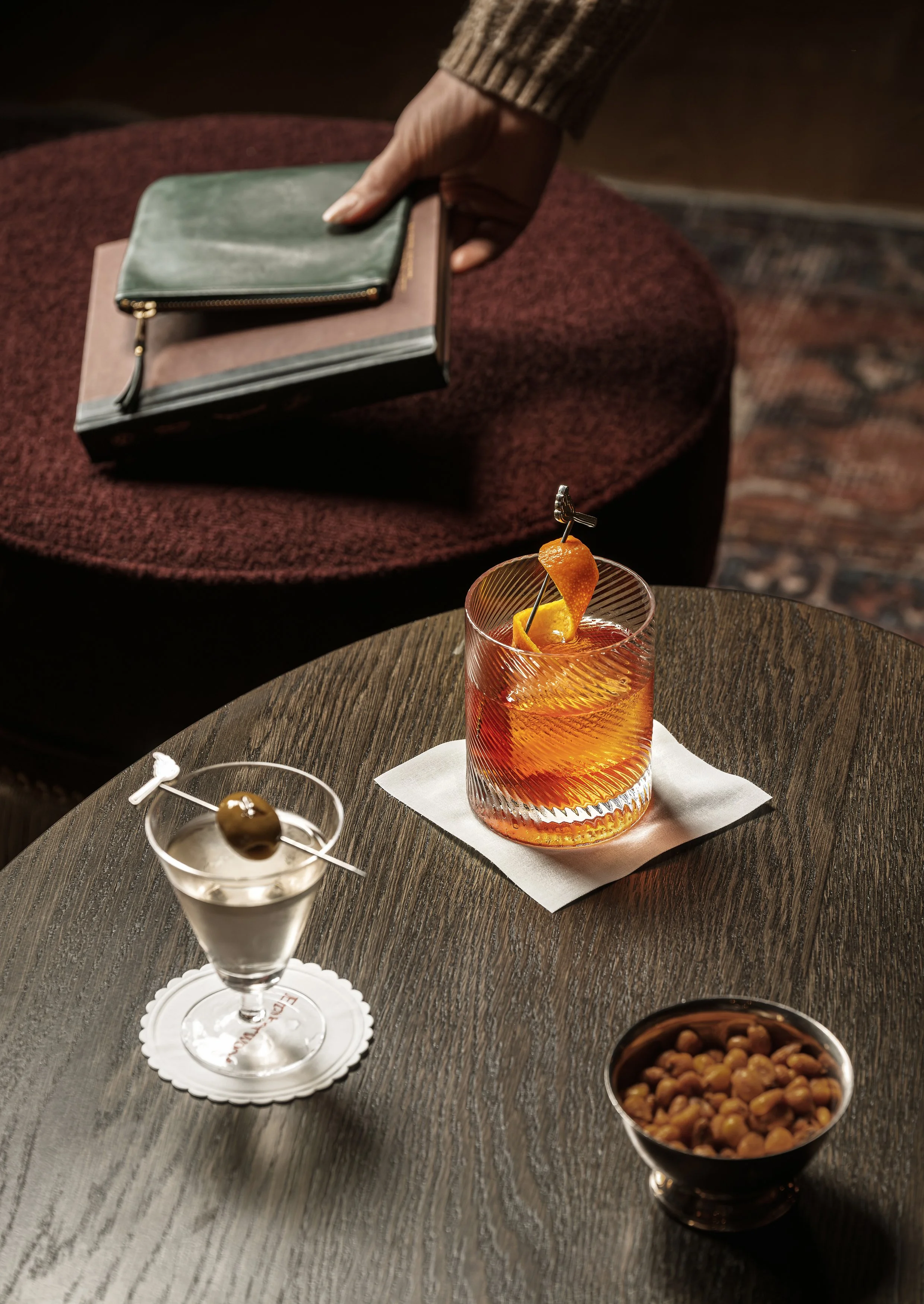 A dark wooden table with a cocktail glass containing an orange drink with an orange twist and a cherry garnish, a smaller glass with a cherry garnish on a paper coaster, and a bowl of mixed nuts. In the background, a person's hand is reaching for a c