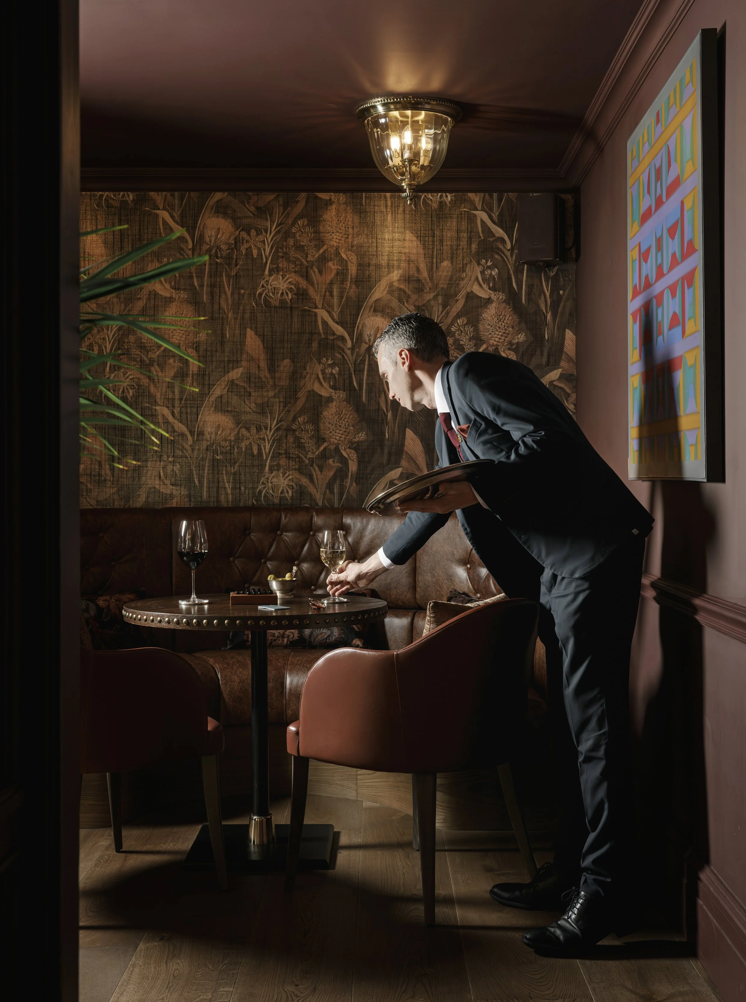 A waiter in a black suit setting a glass of white wine on a table in a dimly lit, elegant restaurant with brown wallpaper, artwork on the wall, and a leather couch.