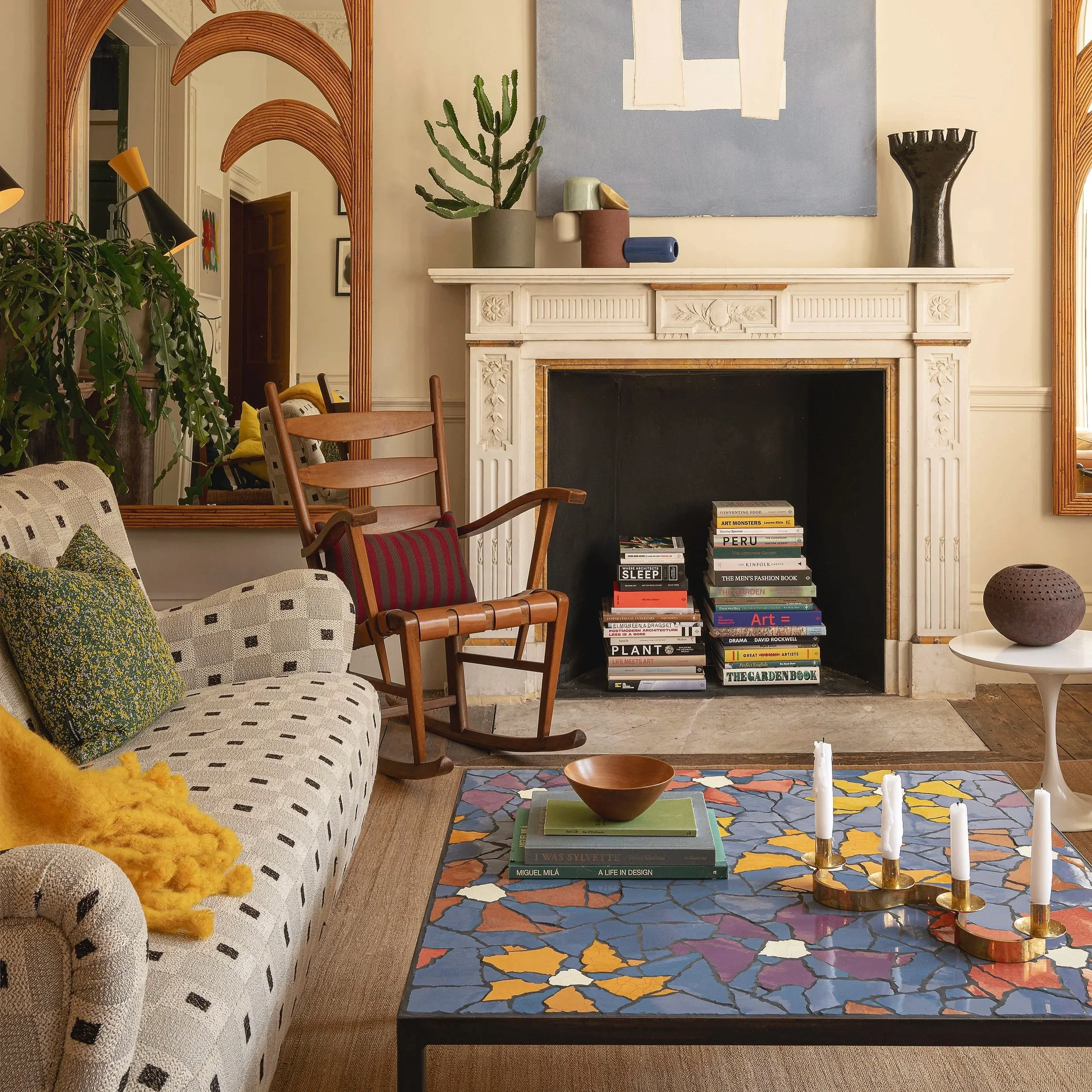 A cozy living room with a black fireplace filled with stacked books, a patterned sofa with green and yellow pillows, a rocking chair with a pillow, a round white side table with a purple vase, and a colorful mosaic coffee table with candles and books. Decor includes a large mirror and various abstract art pieces.