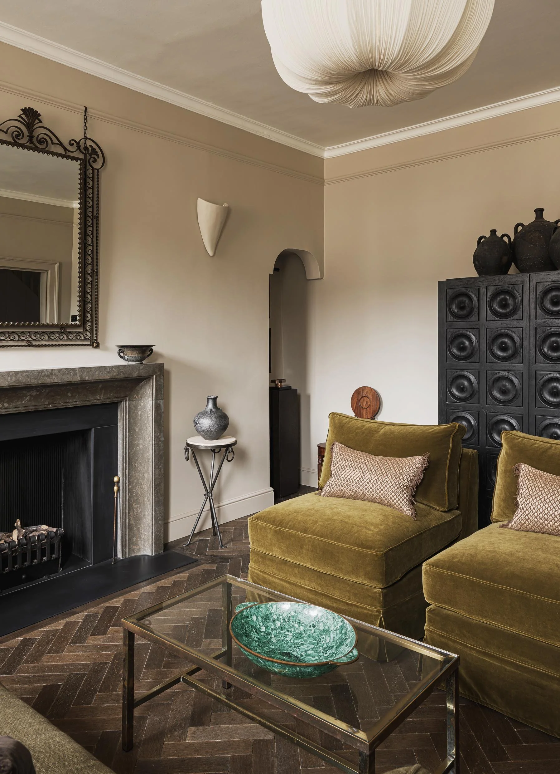 A living room with two olive green velvet armchairs, a glass coffee table with a green ceramic bowl, and a fireplace with a gray stone mantel. Decor includes black and gray vases, a mirror, and a black cabinet with decorative objects, against beige w