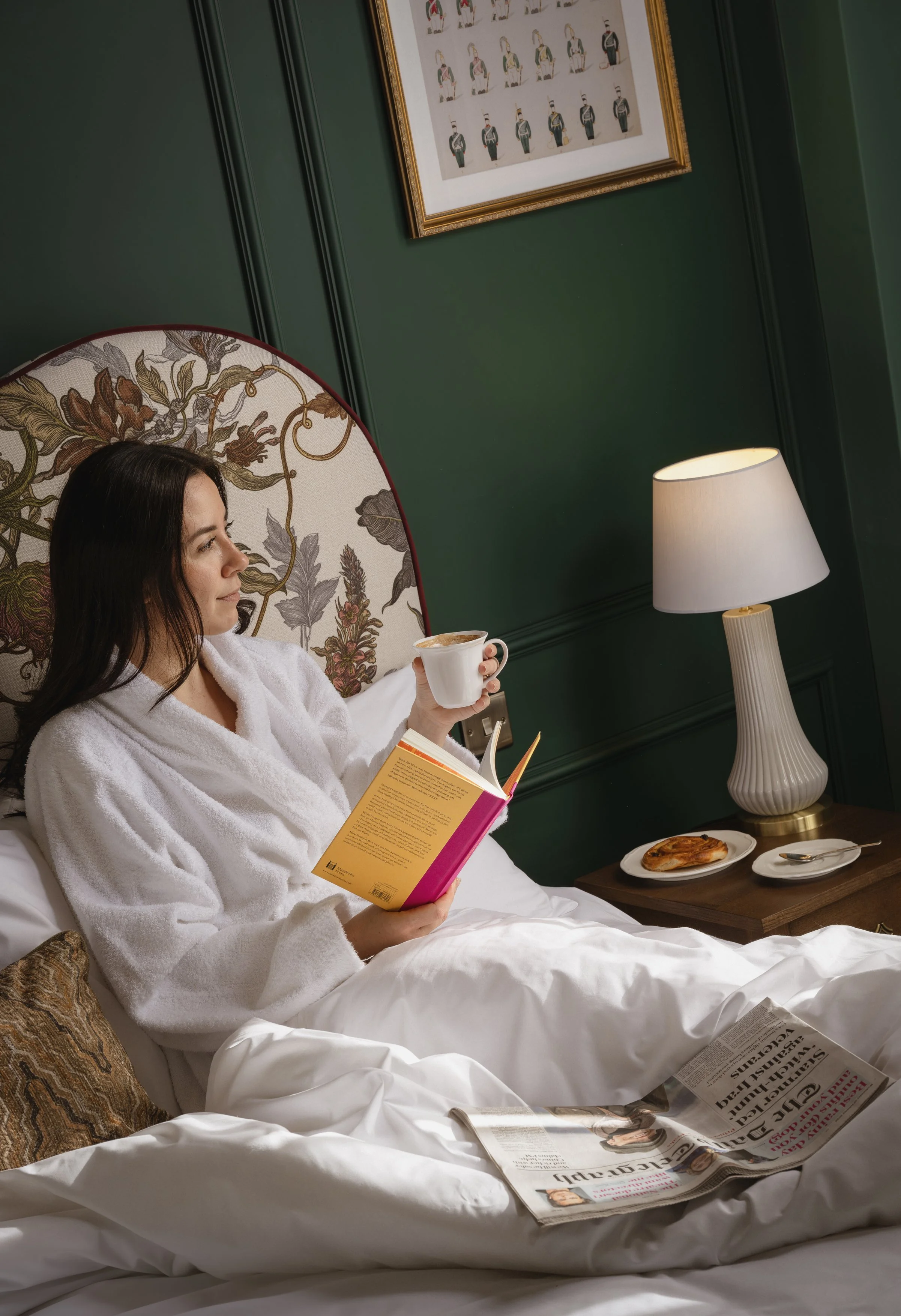 A woman in a white bathrobe sitting in bed, holding a coffee mug and reading a book, with a newspaper on the bed and breakfast plate with a pastry beside her. A bedside lamp and framed artwork are visible in the background.