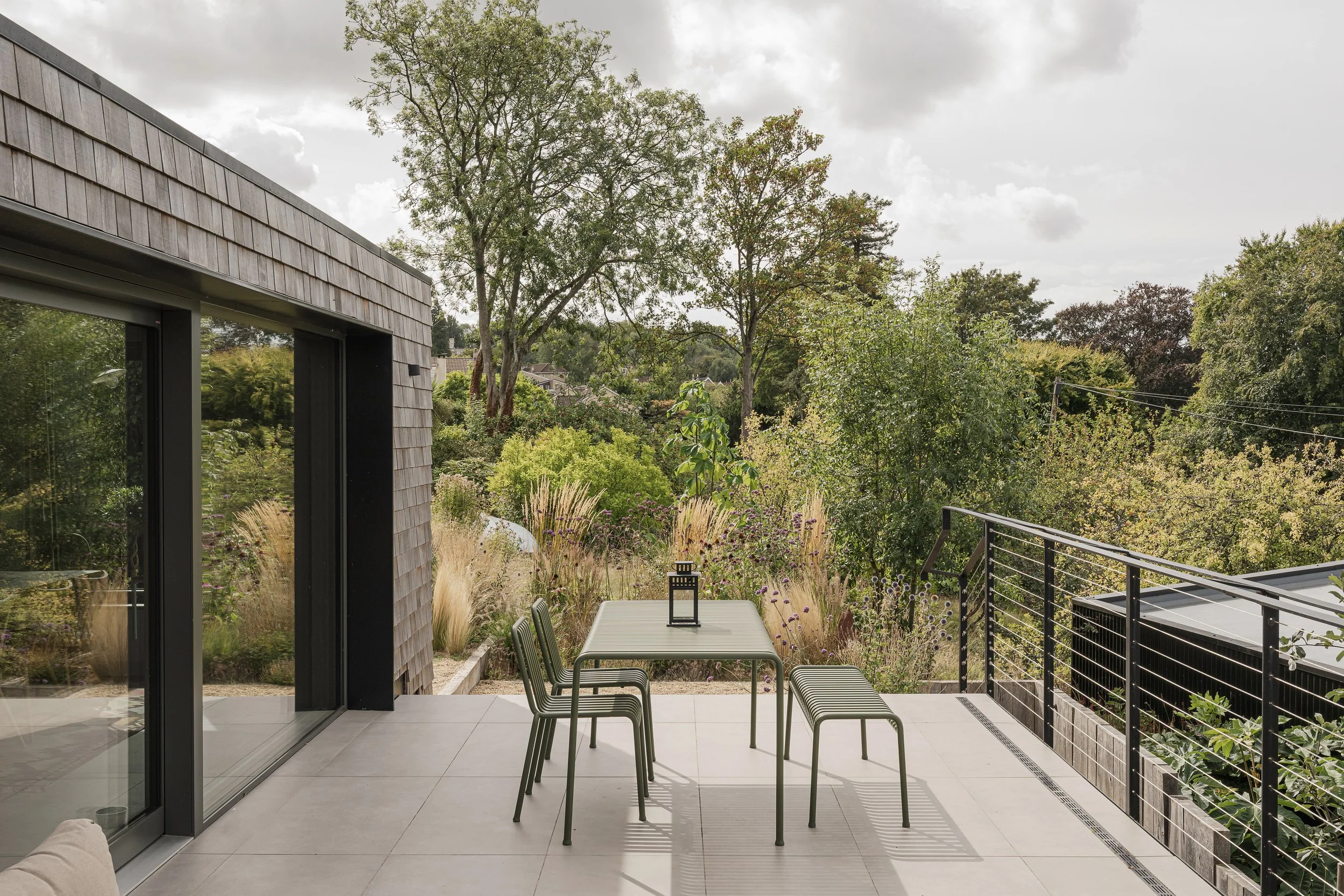 Modern outdoor patio with a gray table, four chairs, and a small lantern, overlooking lush greenery and trees under cloudy sky.