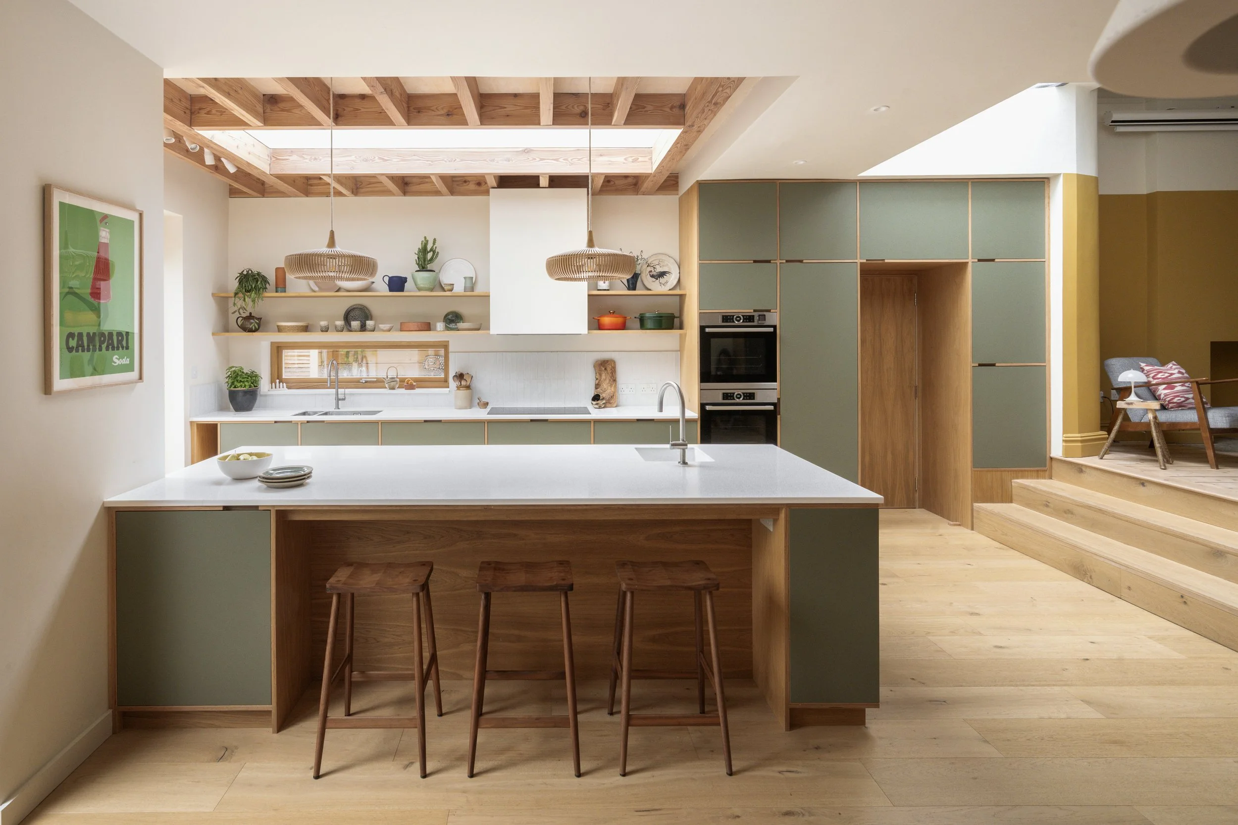 Modern kitchen with wooden cabinets and green accents, island with three wooden stools, open shelving with plants and dishes, and a skylight ceiling.