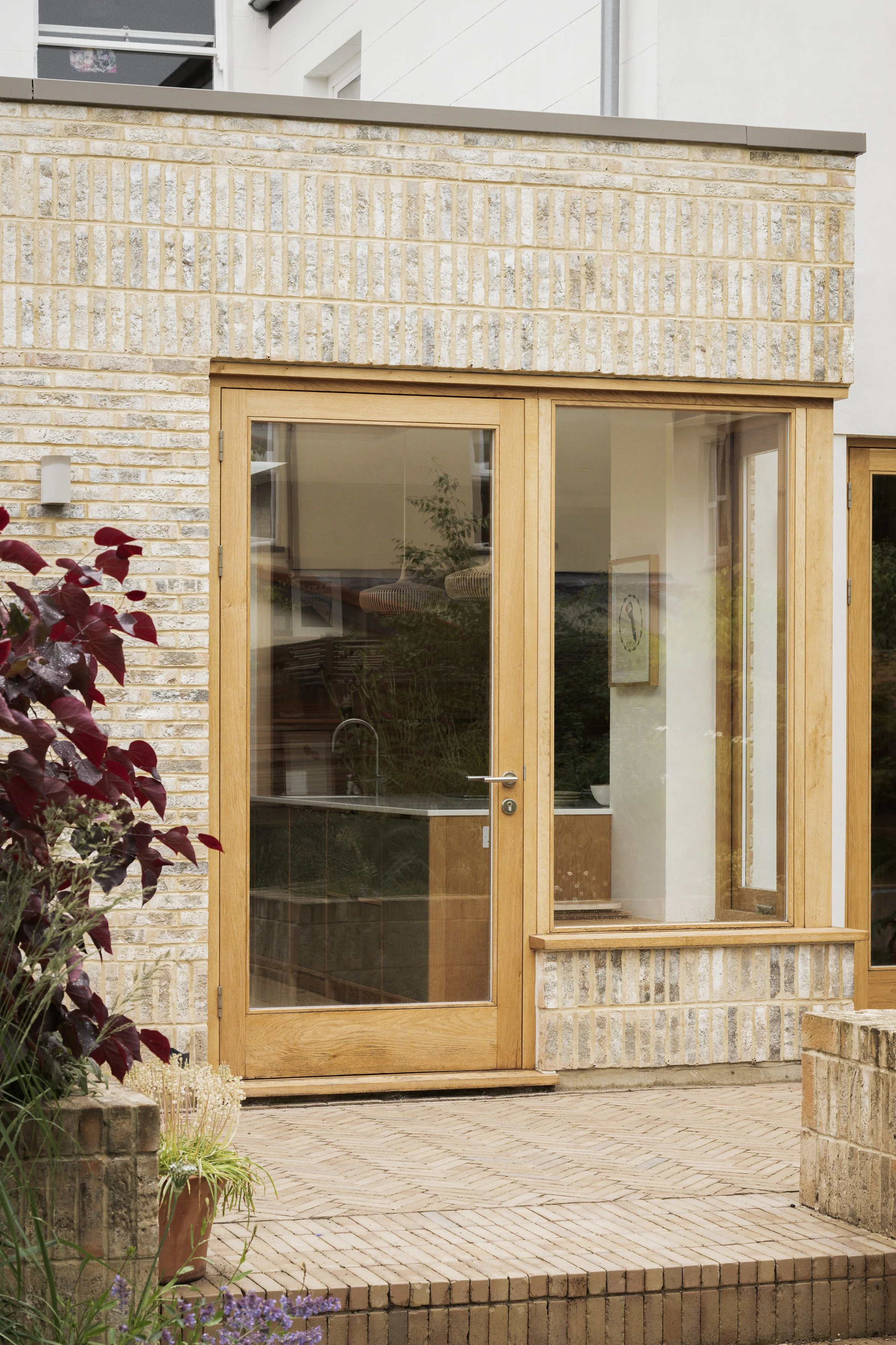 Exterior view of a building with a wooden framed glass door and window, brick wall, potted plants, and a brick patio.