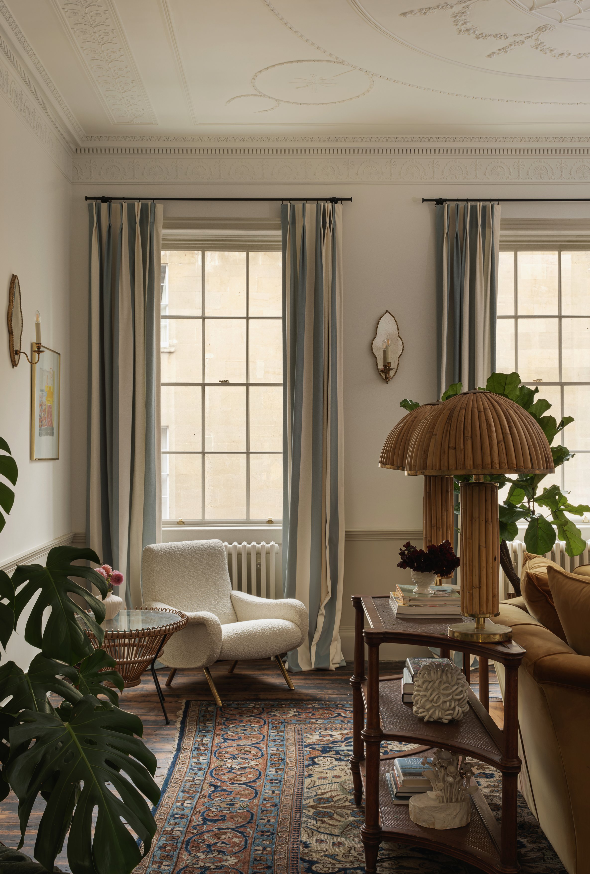Bright living room with two large windows framed by blue and white striped curtains, a white armchair, a side table with a pink flower bouquet, a wooden side table with books and a ceramic decorative piece, a large wooden lampshade, and various plant